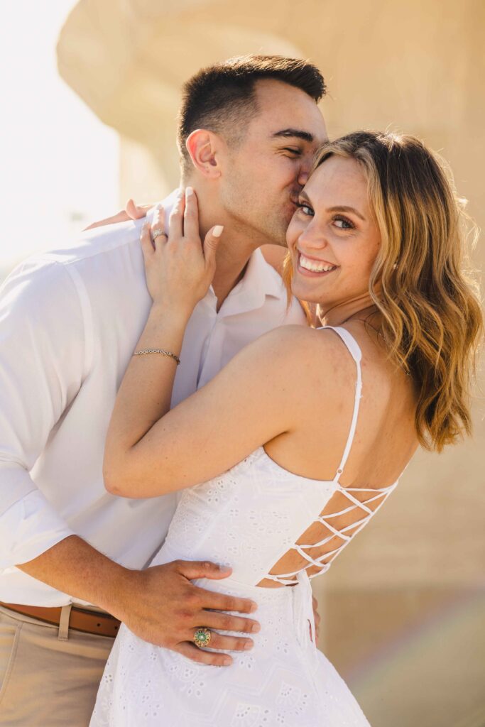 Man kissing his fiancee's head as she smiles to the camera.