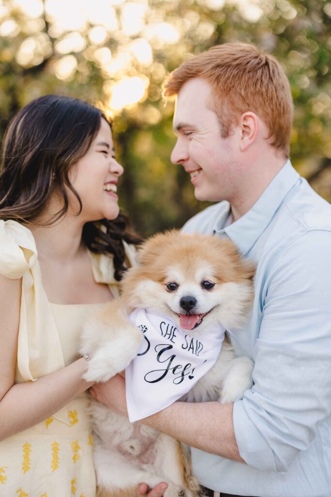 A couple holding their dog smiling. The dog is wearing a bandana that says "she said yes!" 