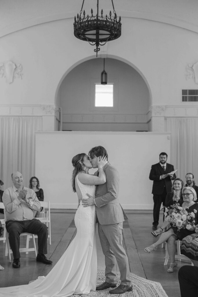 Bride and groom kissing at their wedding reception with guests clapping in the background.