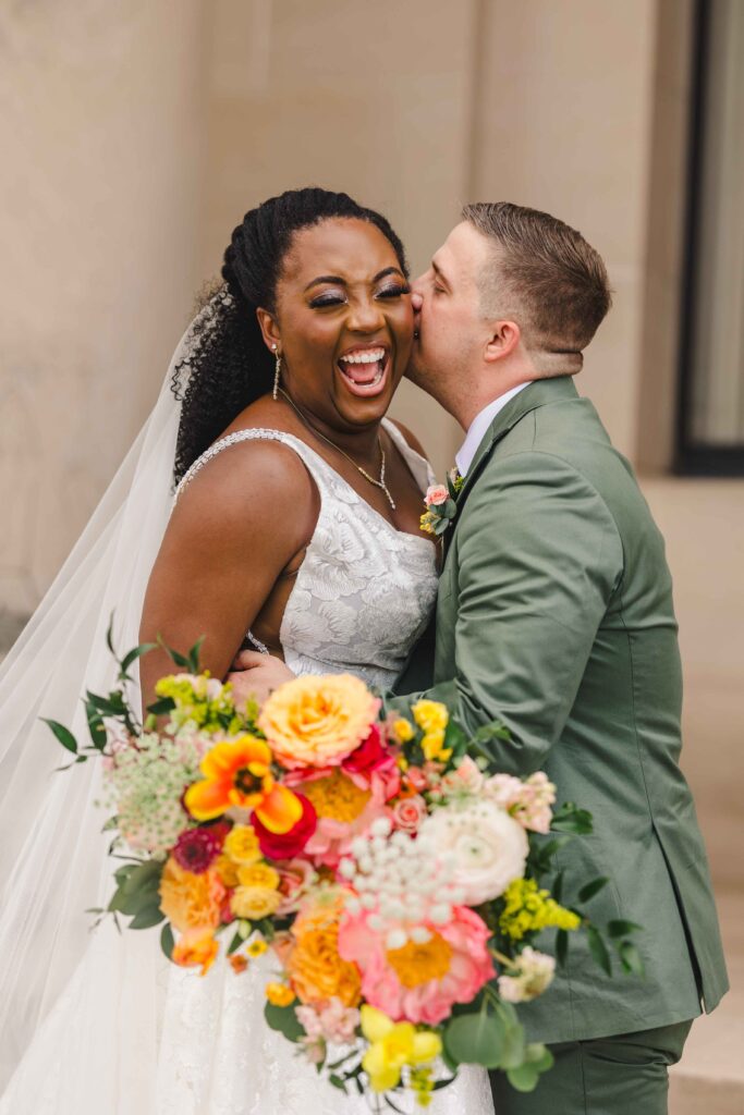 Groom kissing his brides cheek as she smiles playfully. 