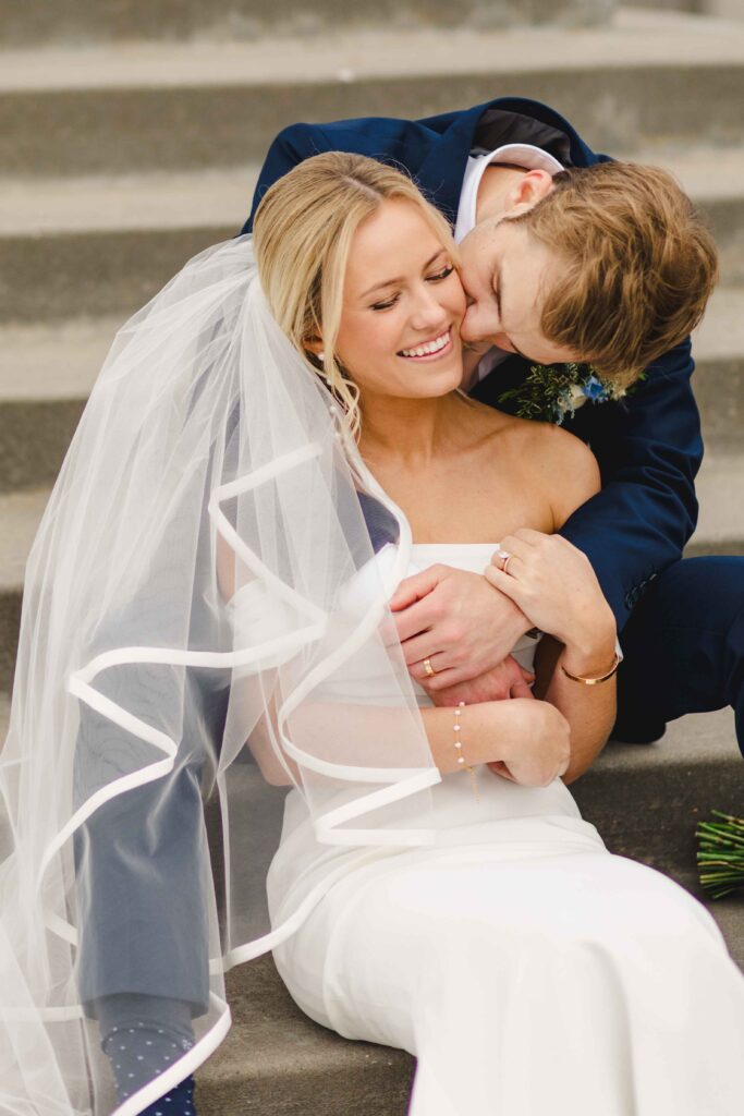 Bride and groom sitting on steps. The groom is kissing the bride on the cheek.