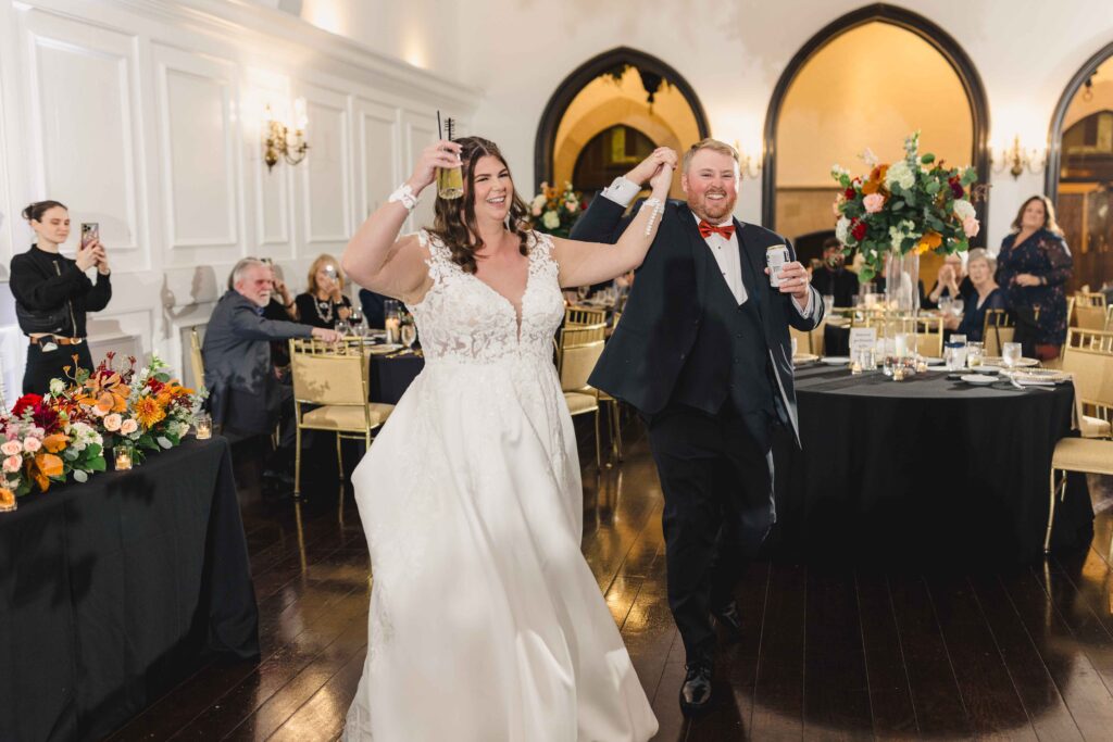 The newlyweds holding up drinks and walking into their Oakwood Country Club wedding reception as guests cheer. 