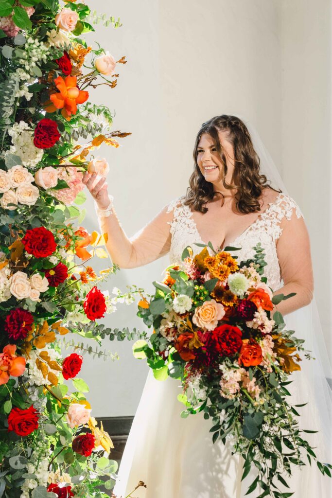 Bride by the flower arch altar holding a large flower bouquet. 