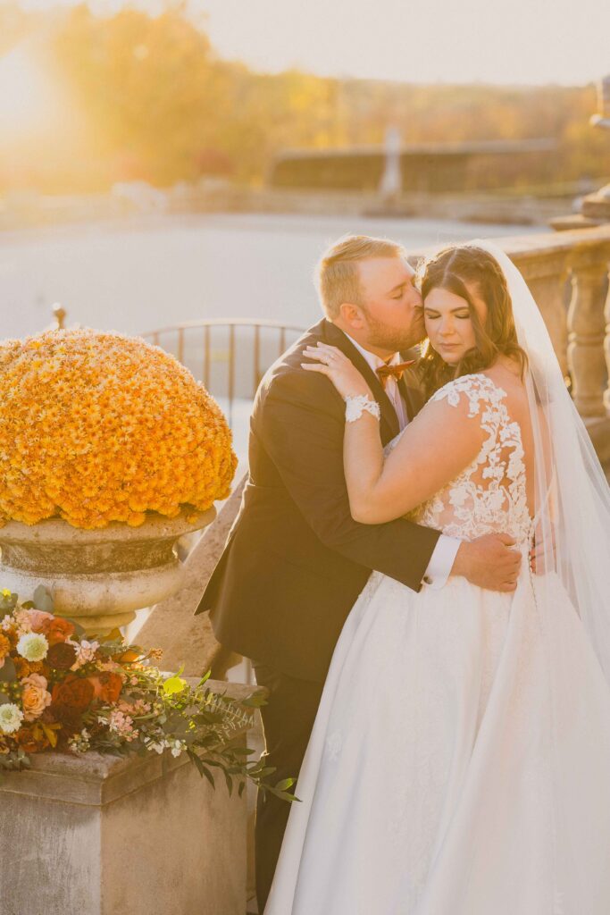 Groom kissing his wife's cheek as the sun sets behind them. 