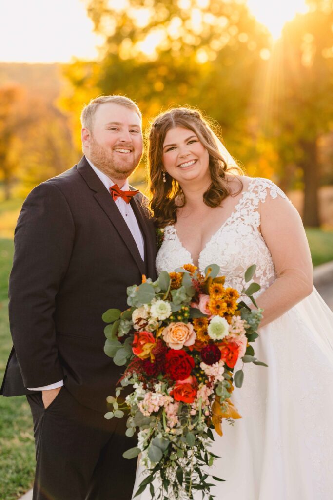 Bride and groom smiling while holding a large bouquet as the sunsets. 