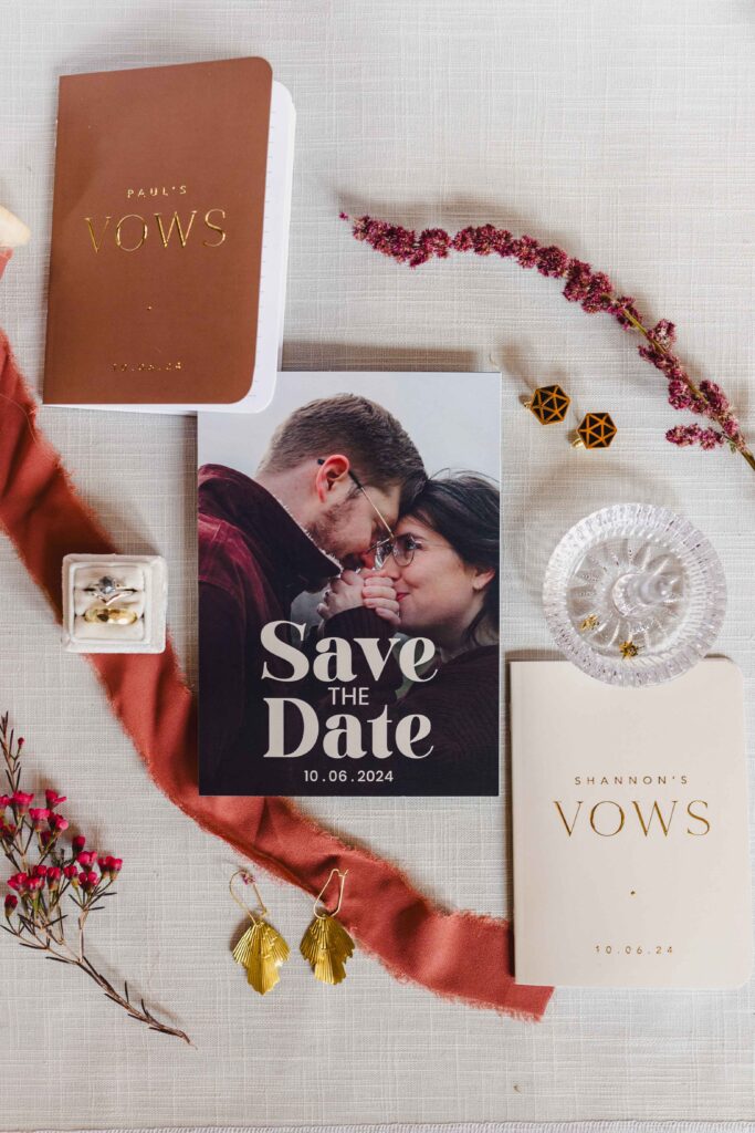 Wedding details of the save the date card, vow books, and rings. 