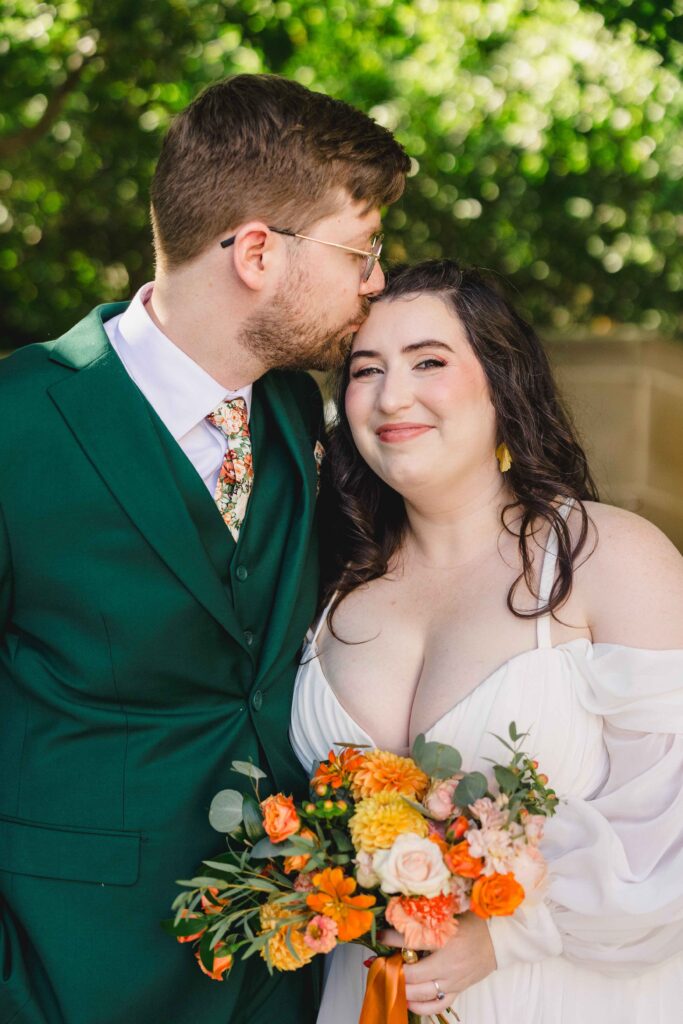 Groom kissing the bride's head as she smiles and holds a vibrant wedding bouquet. 