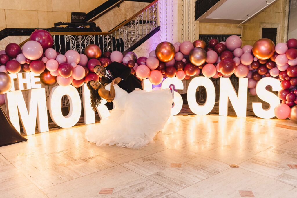 Groom dipping his bride in front of a light up sign that says "The Morrisons" with colorful balloons in the background.