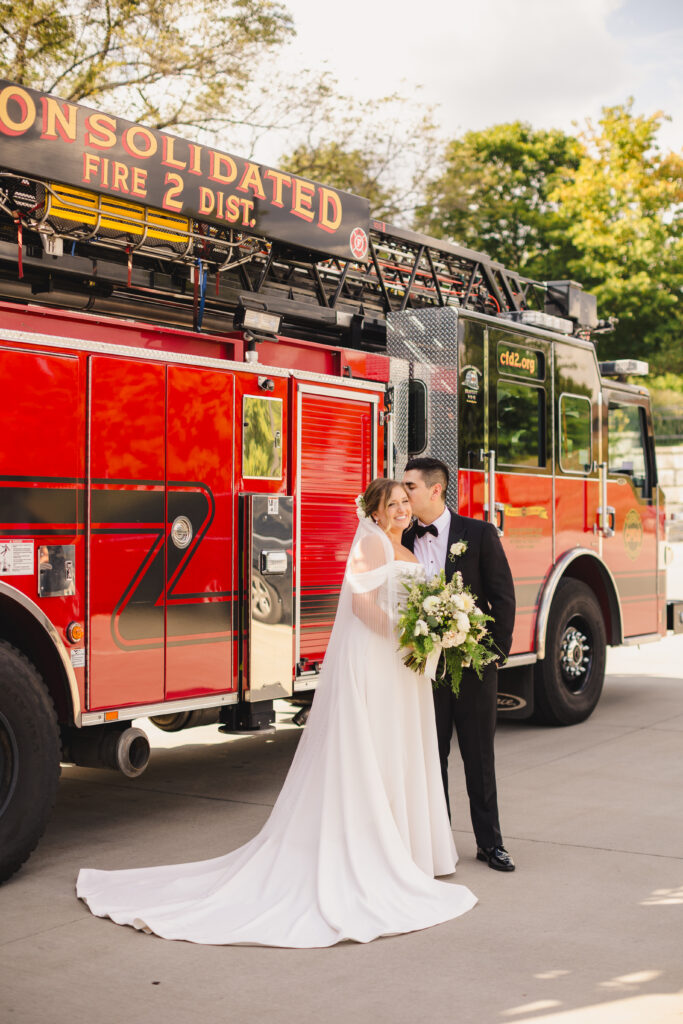 Groom kissing his wife's head while standing by a fire truck.