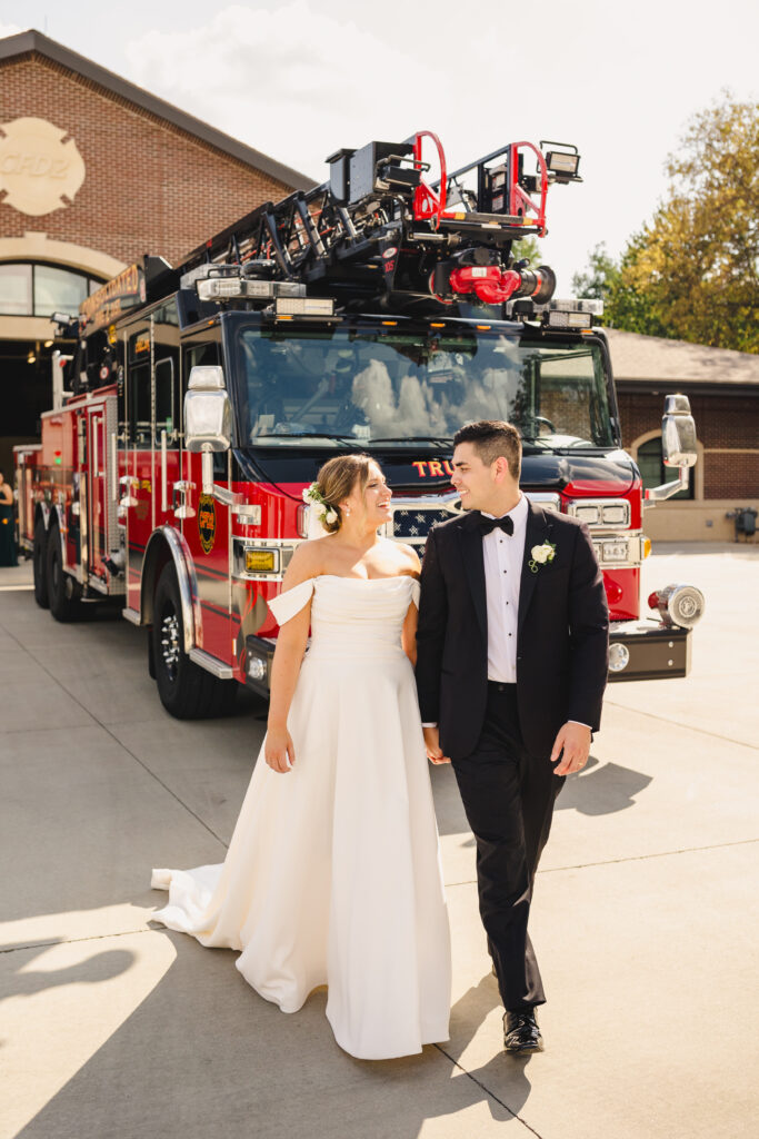 Bride and groom walking with a fire truck behind them.