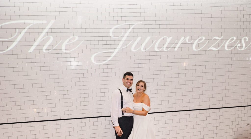 The newlyweds standing outside The Guild KC by a white brick wall displaying "The Juarezes"