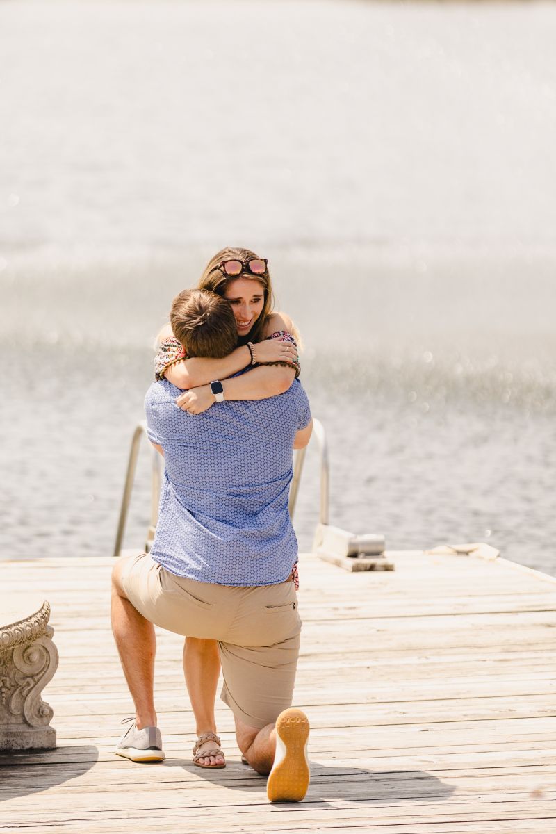 a man down his knees proposing to his partner next to the water on a dock and his partner is hugging him