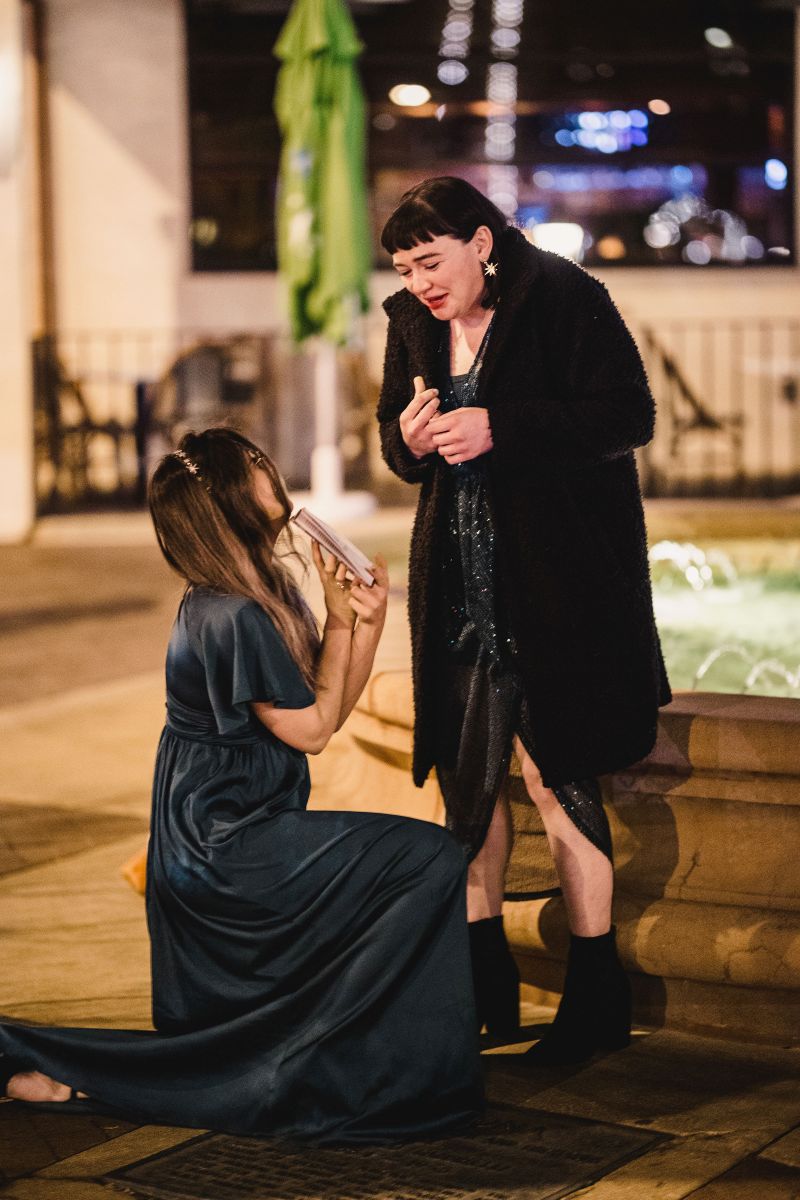 a woman surprising her partner by proposing to her next to a fountain