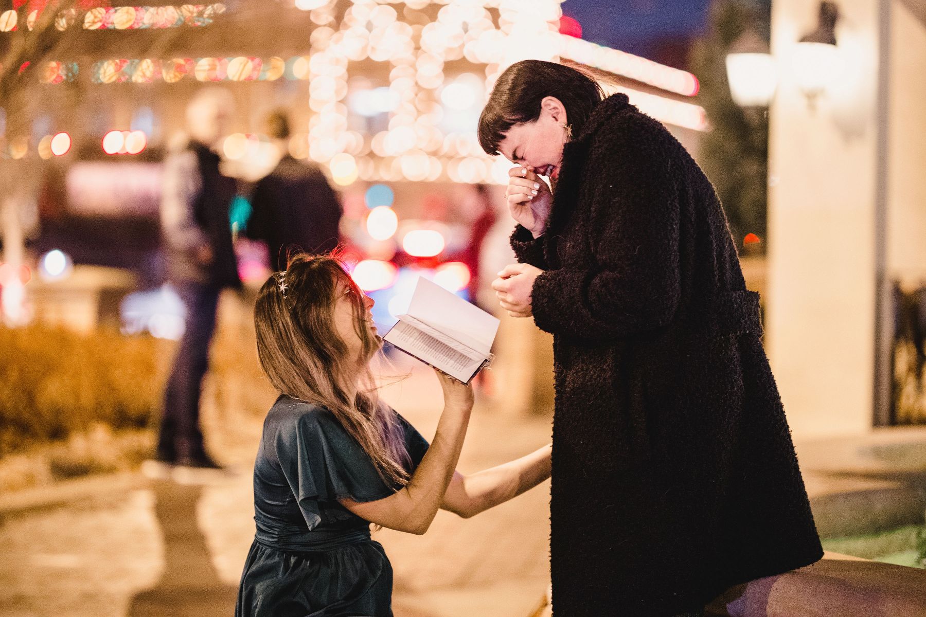 a woman down on her knees with a book in her hand asking her partner to marry her and her partner is crying