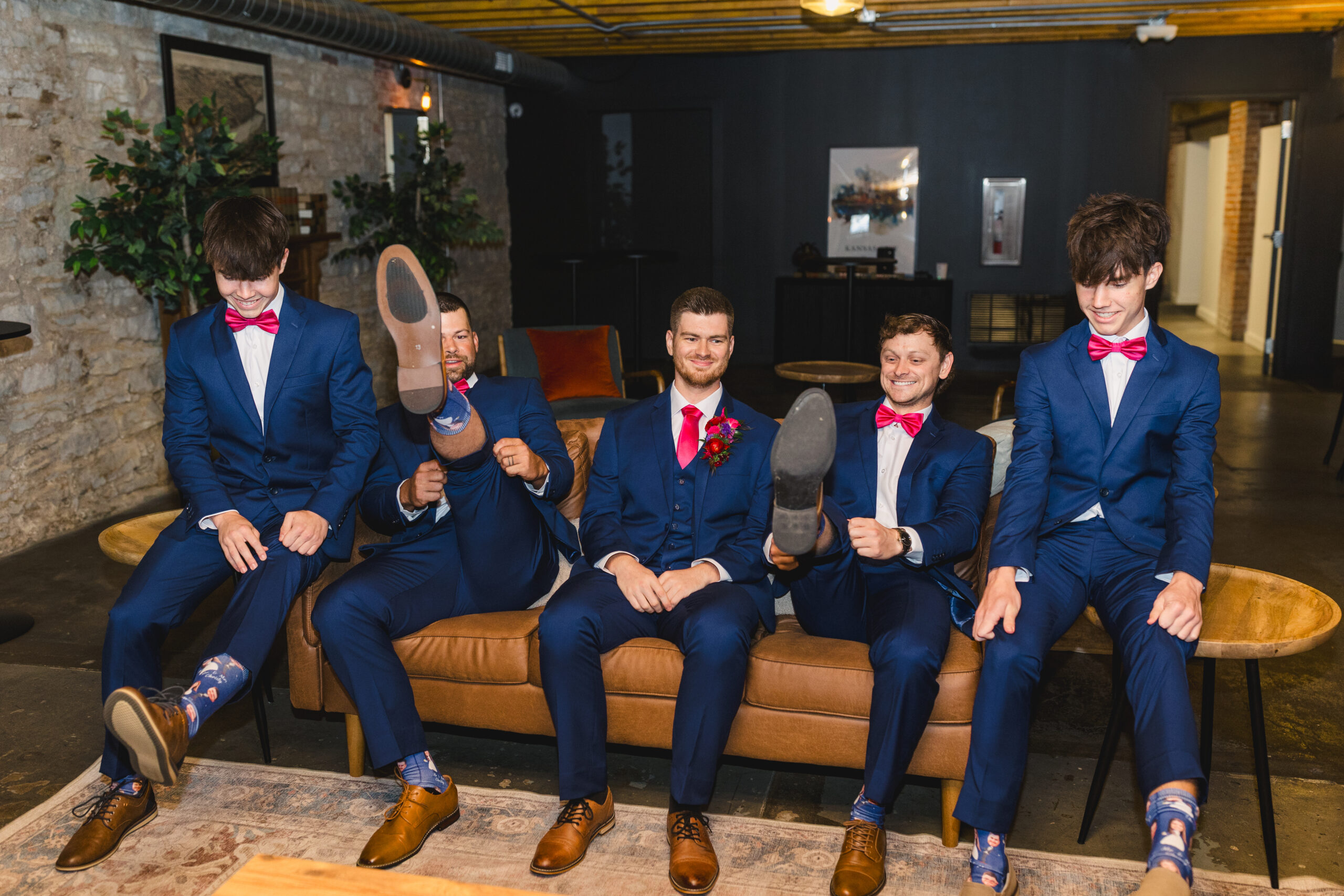 a group of groomsmen sitting on a leather couch all wearing matching blue suits and pink bowties with the groom wearing a pink tie 