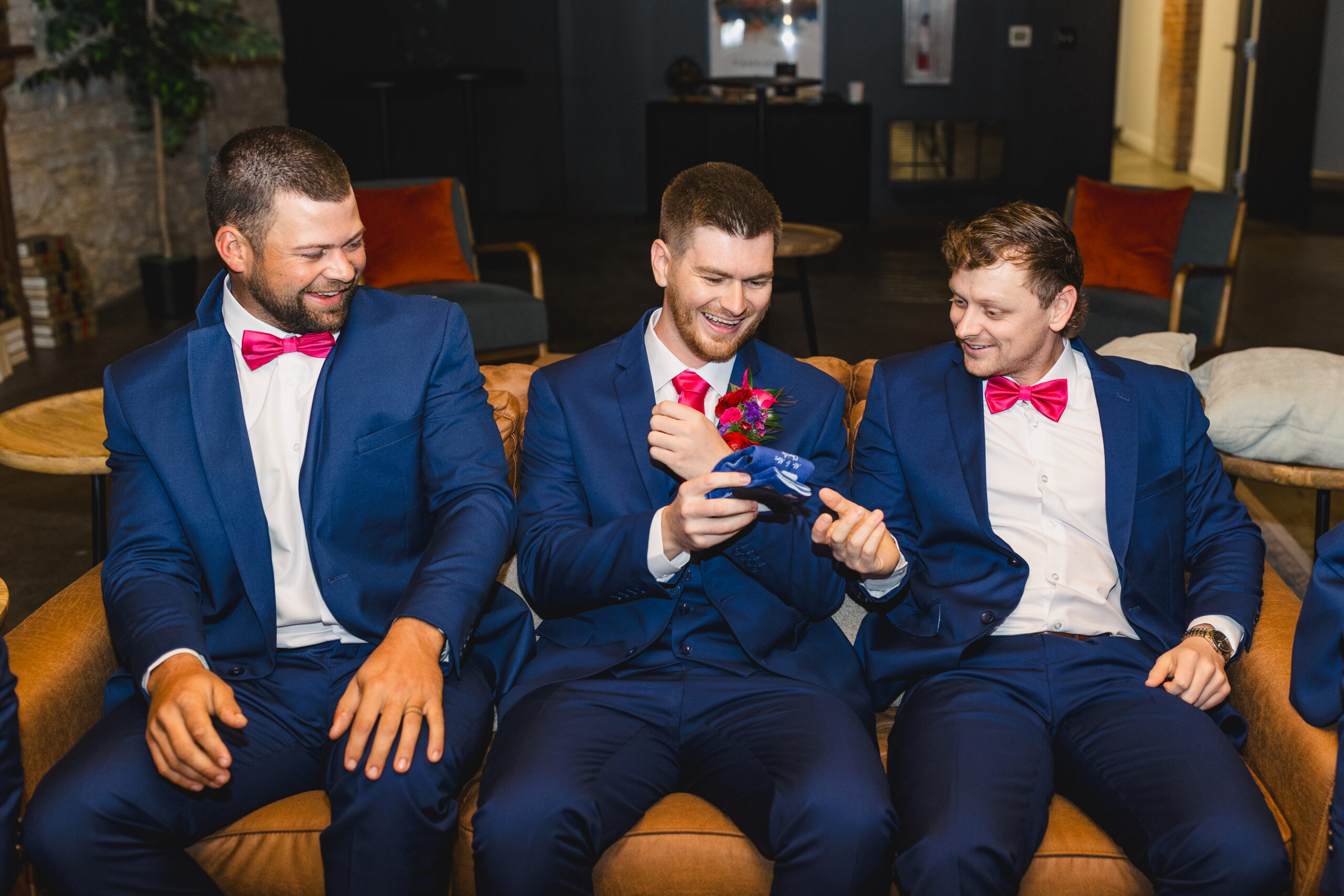 two groomsmen giving the groom a pair of socks on the day of his wedding they are all laughing together 