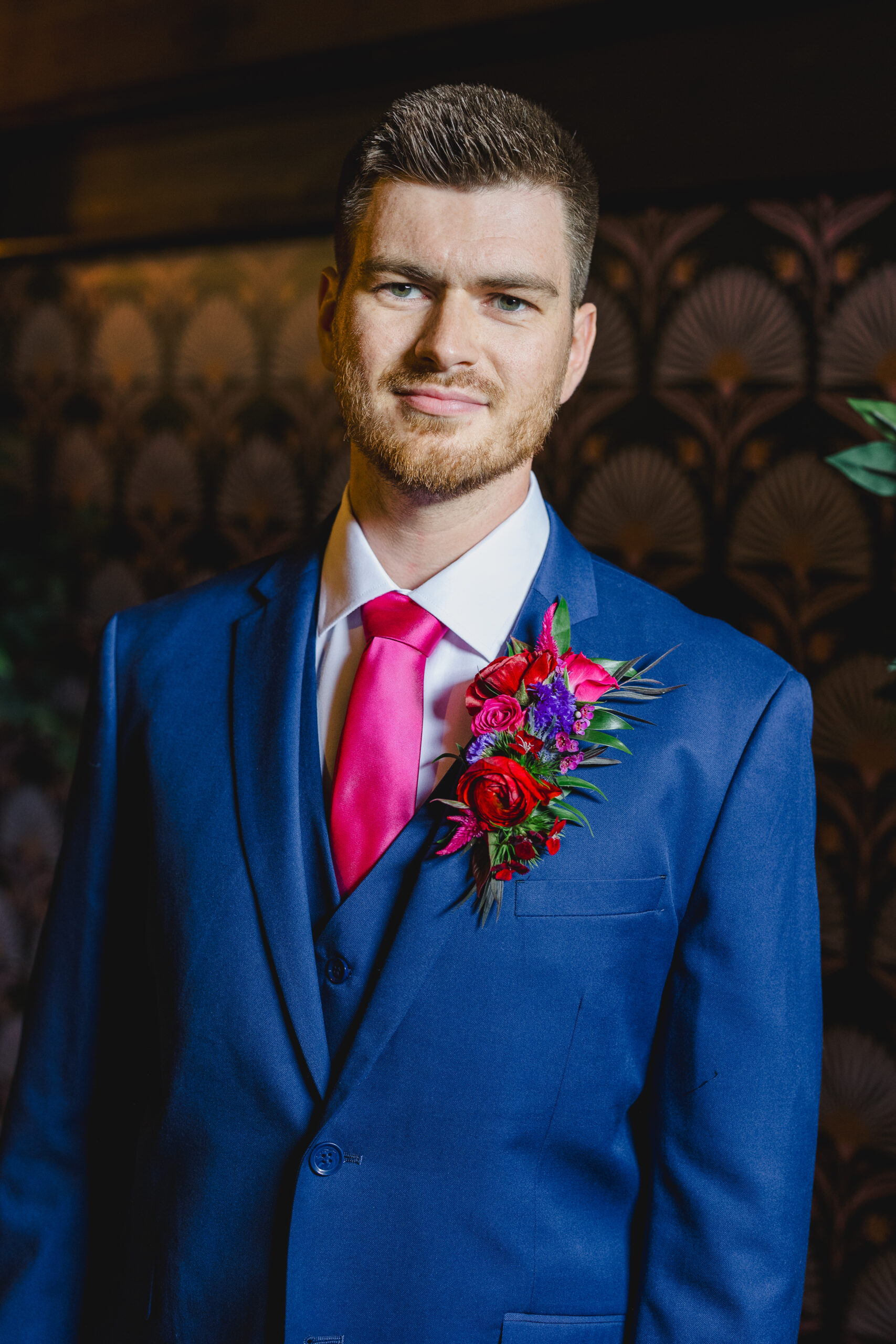 a man wearing a blue suit and pink tie softly smiling at the camera and he is wearing a blue and pink boutonniere 