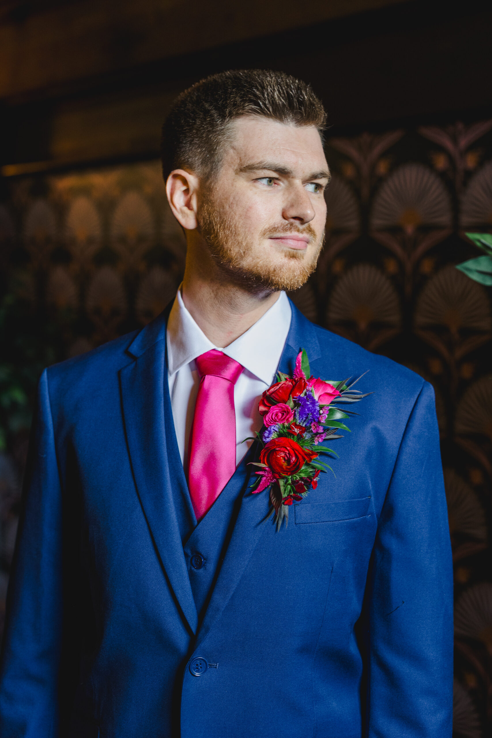 a man wearing a blue suit and pink tie softly smiling with his head tilted to the side and he is wearing a blue and pink boutonniere 