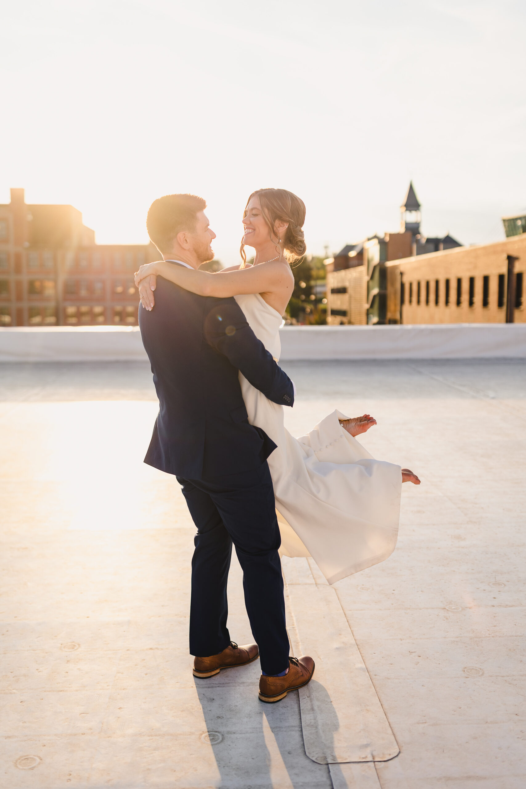 a man picking up his partner they are taking golden hour portraits on the rooftop of union in kansas city the skyline is behind them 