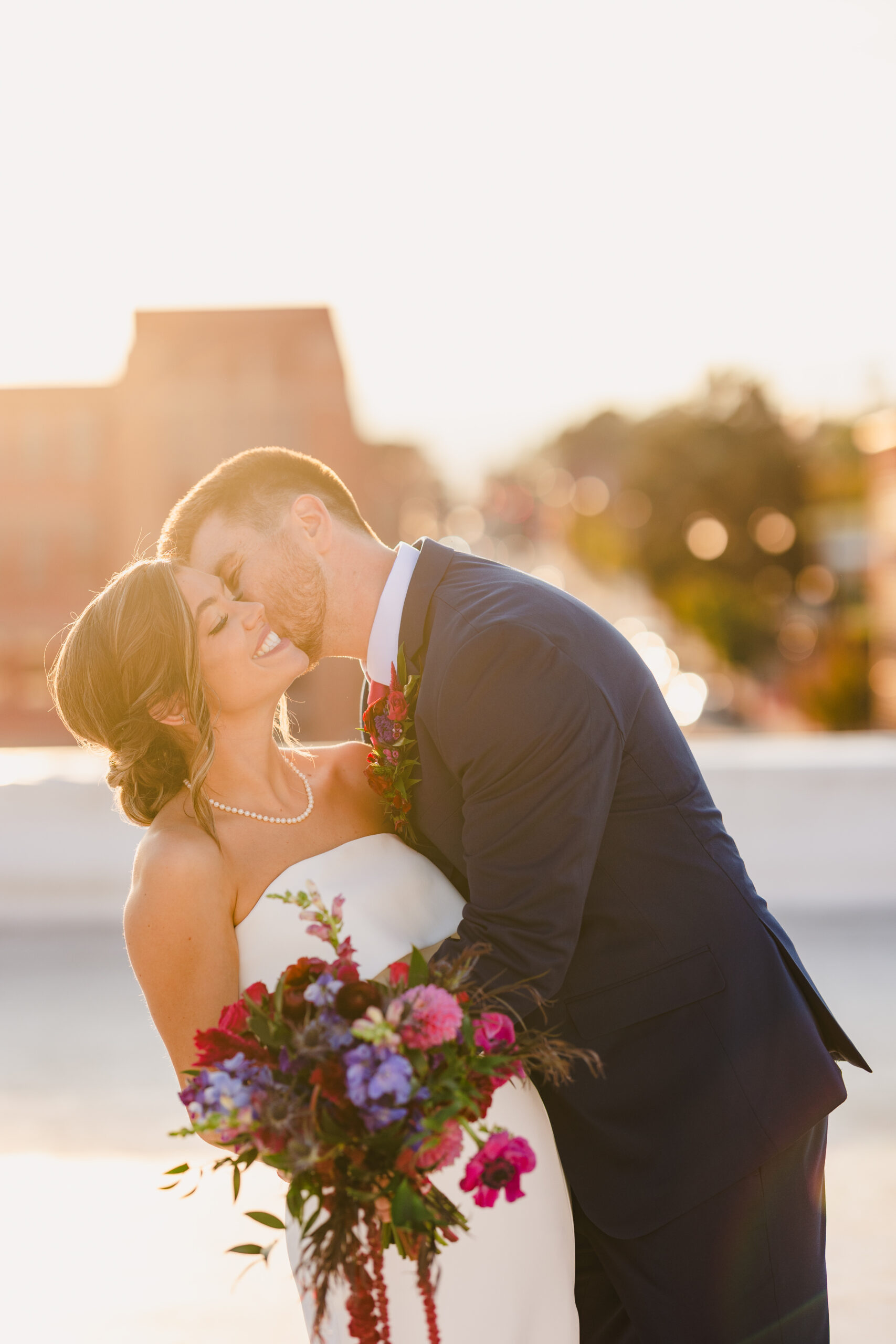 a couple taking golden hour photos on the rooftop of union in kansas city with the kansas city skyline behind them the man is hugging his partner and giving her a kiss on the cheek