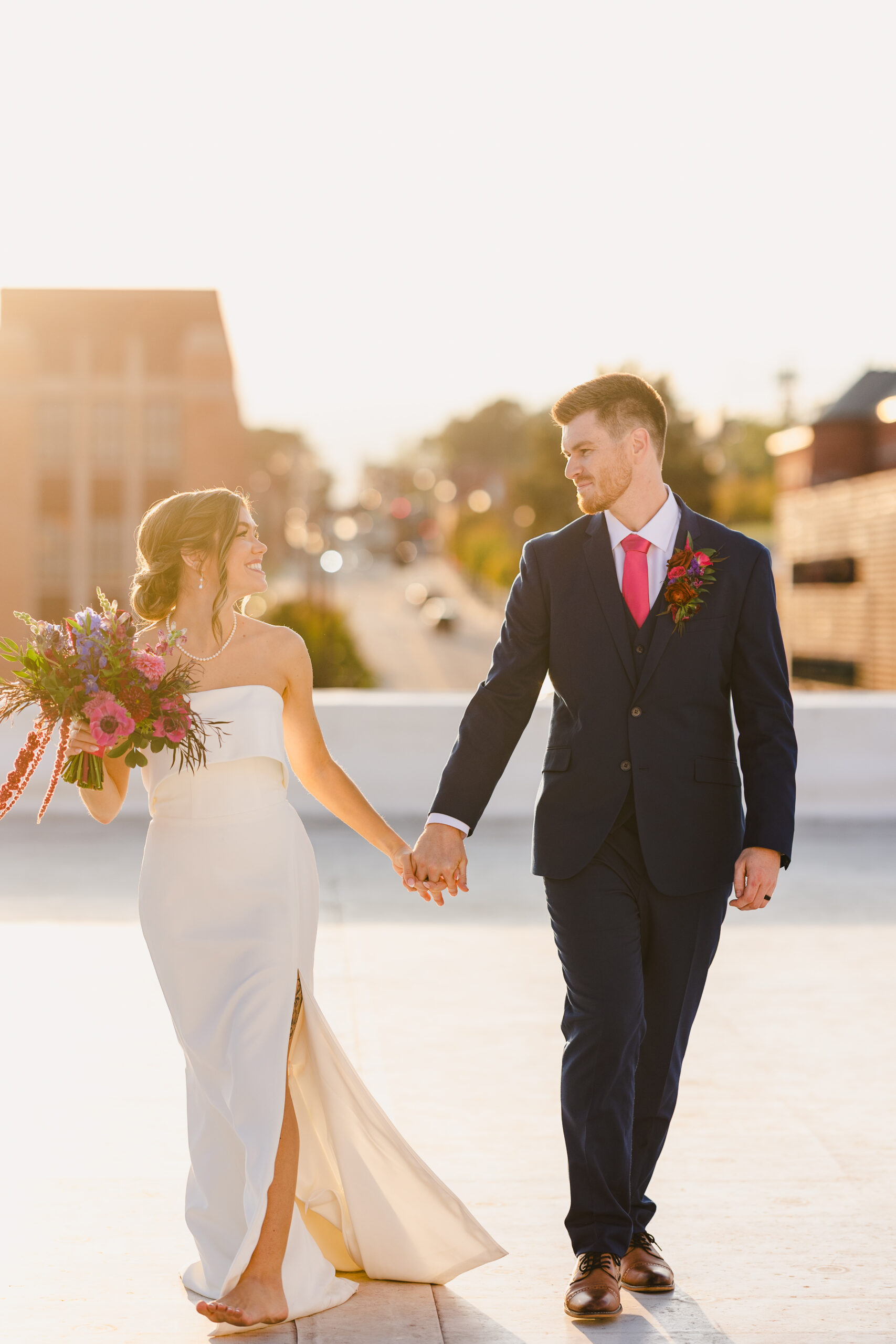 a couple holding hands and smiling at each other they are taking golden hour portraits on the rooftop of union in kansas city 