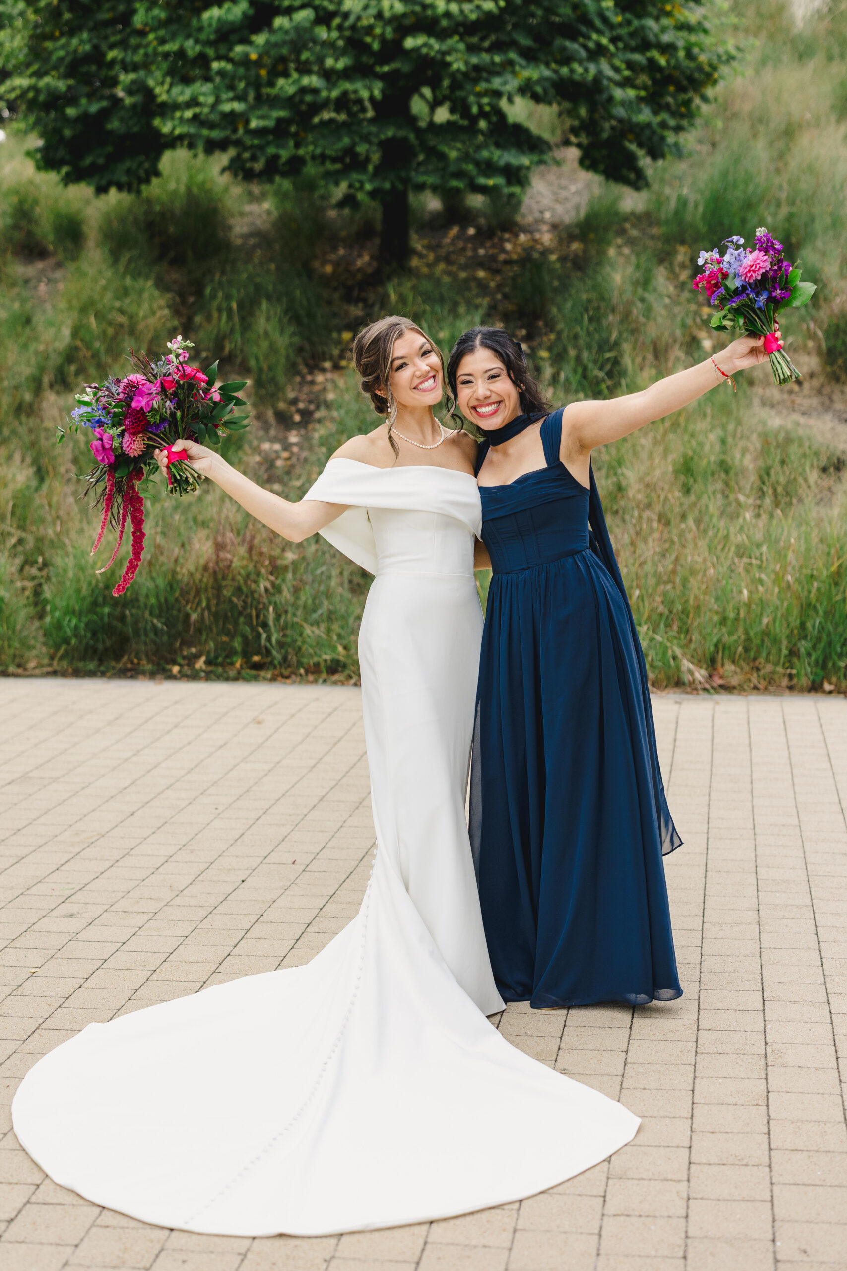a bride posing with a bridesmaid they are both holding up their bouquets in the air and hugging each other 