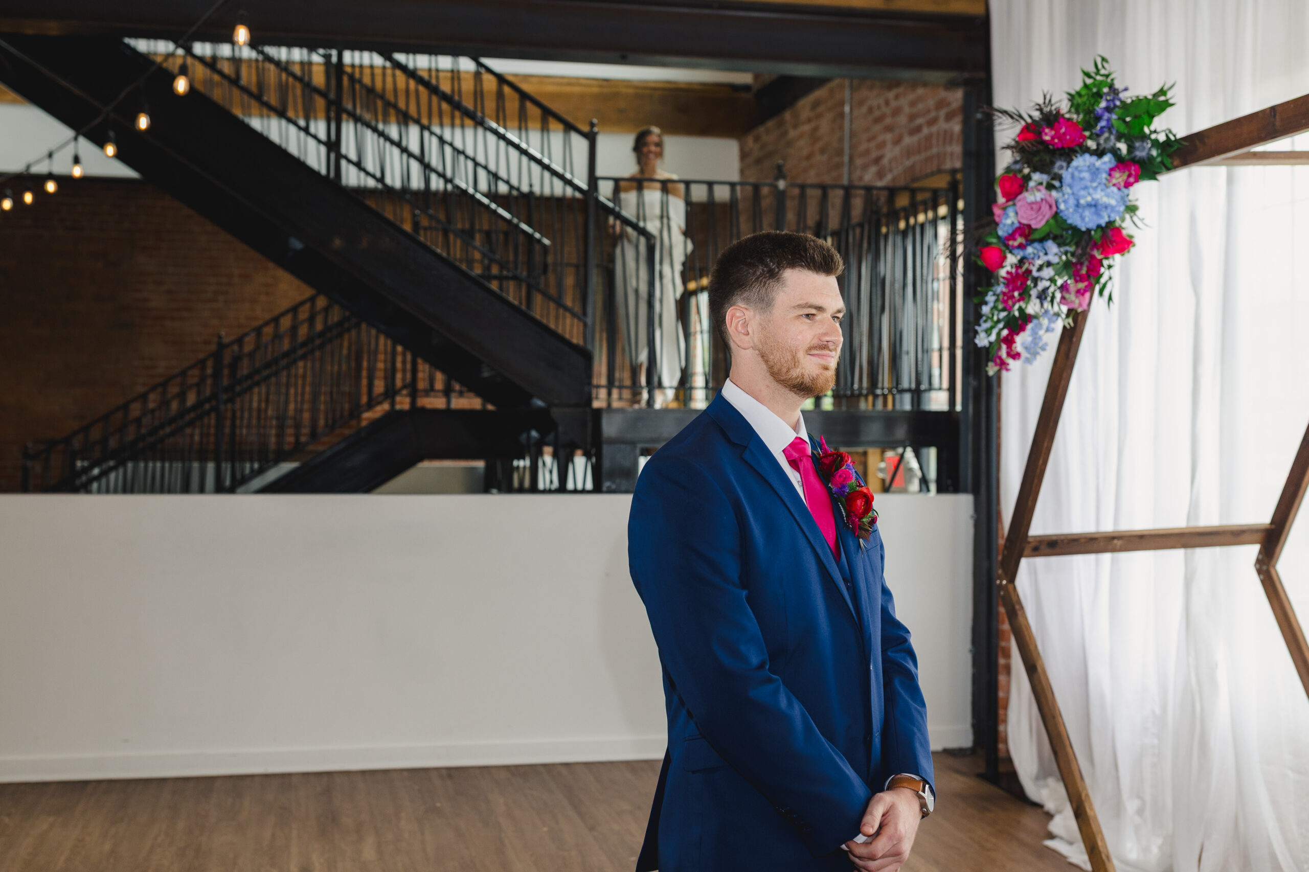 a groom waiting to share a first look with his partner who is coming down the staircase behind him 