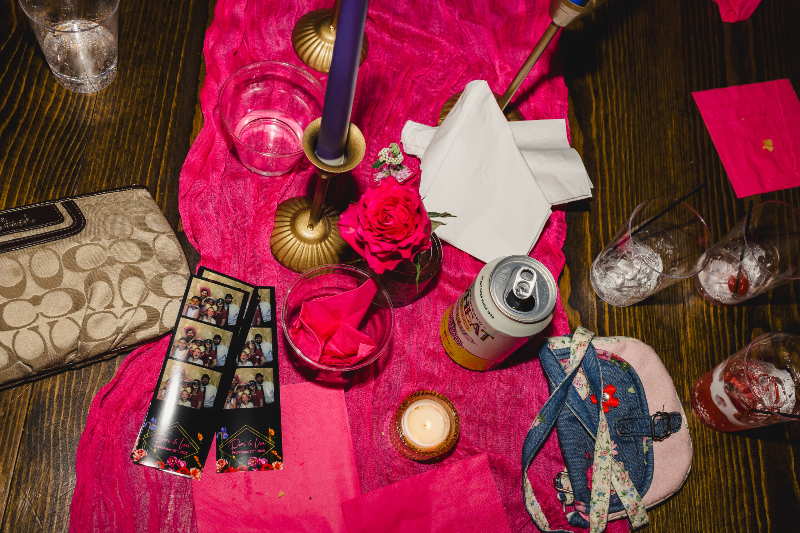 a wooden table on top of it is a pink tablecloth with photos from a photobooth, candles, purses, and drinks 