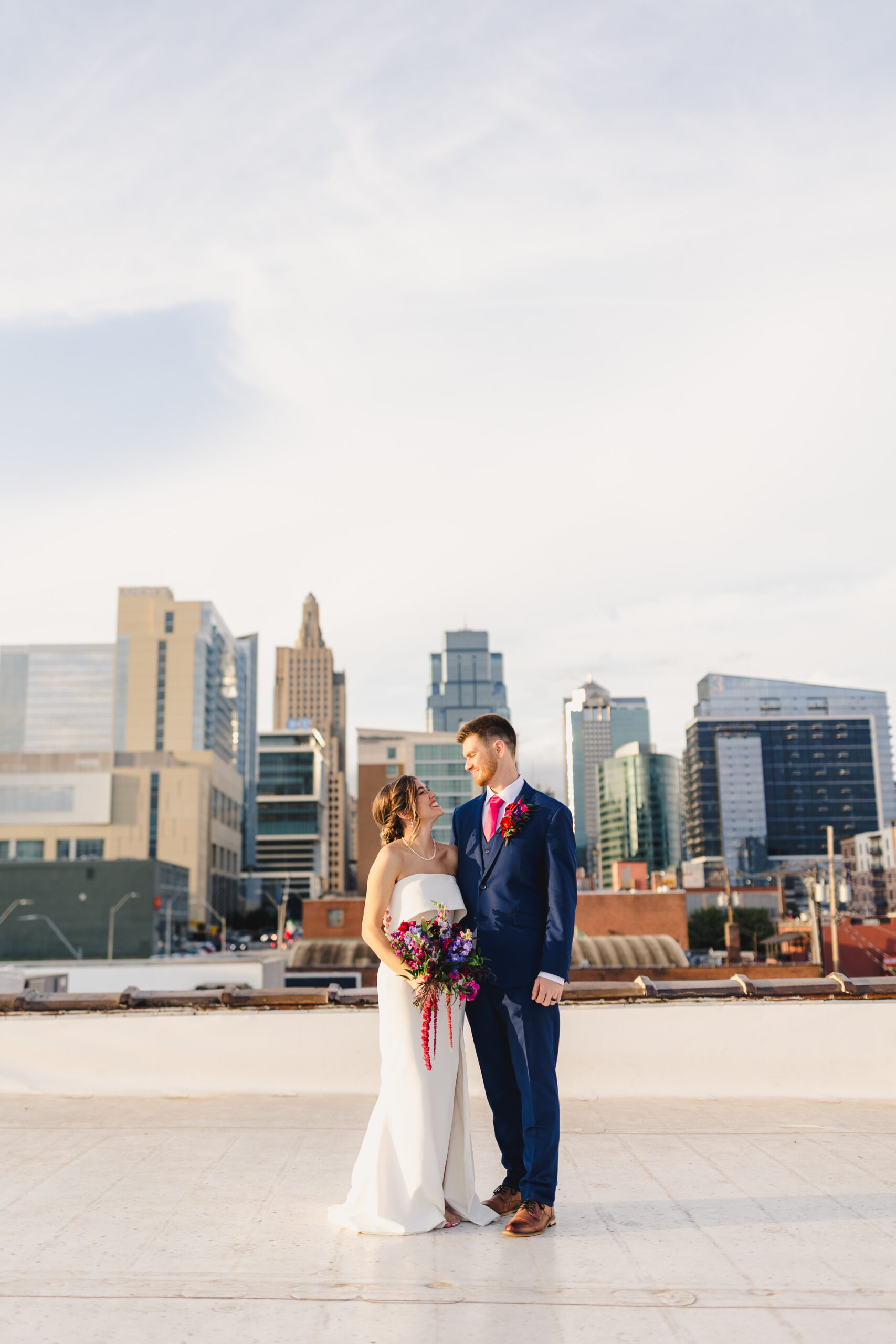 a couple taking golden hour portraits with the kansas city skyline behind them they are both looking at one anotehr and smiling 