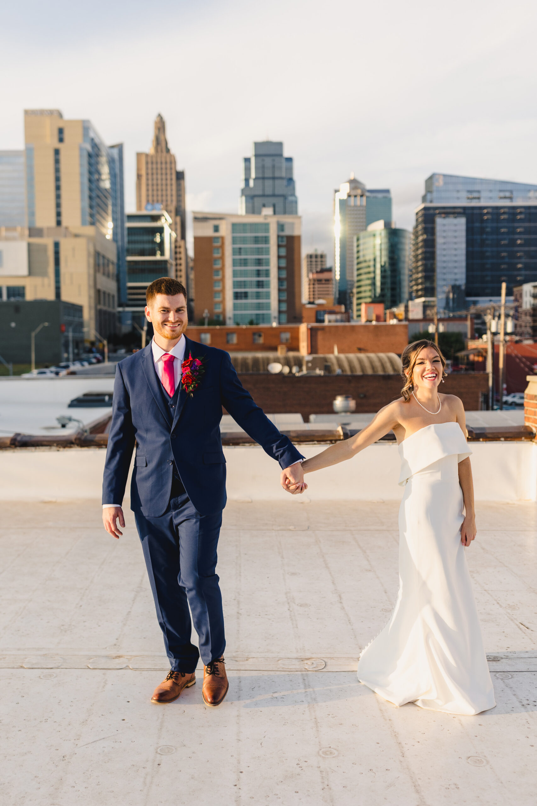 a couple taking golden hour wedding portraits and holding hands with the kansas city skyline behind them 