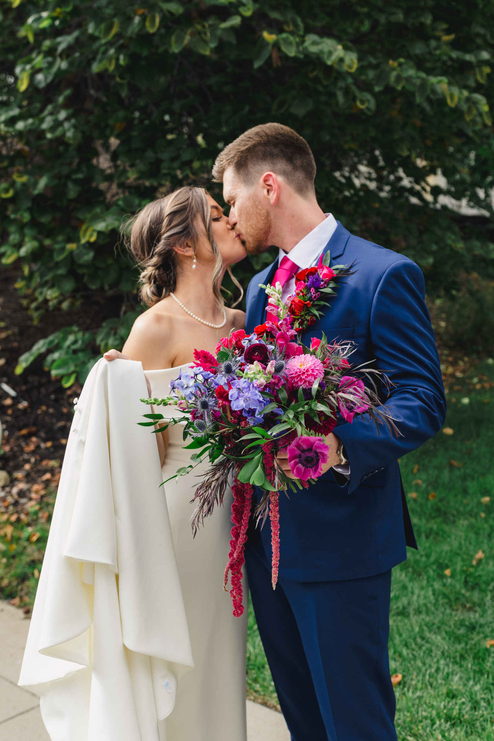 a couple kissing each other next to a tree taking wedding portraits the man is wearing a blue suit and holding a colorful and vibrant floral arrangment consisting of blue and pink flowers 