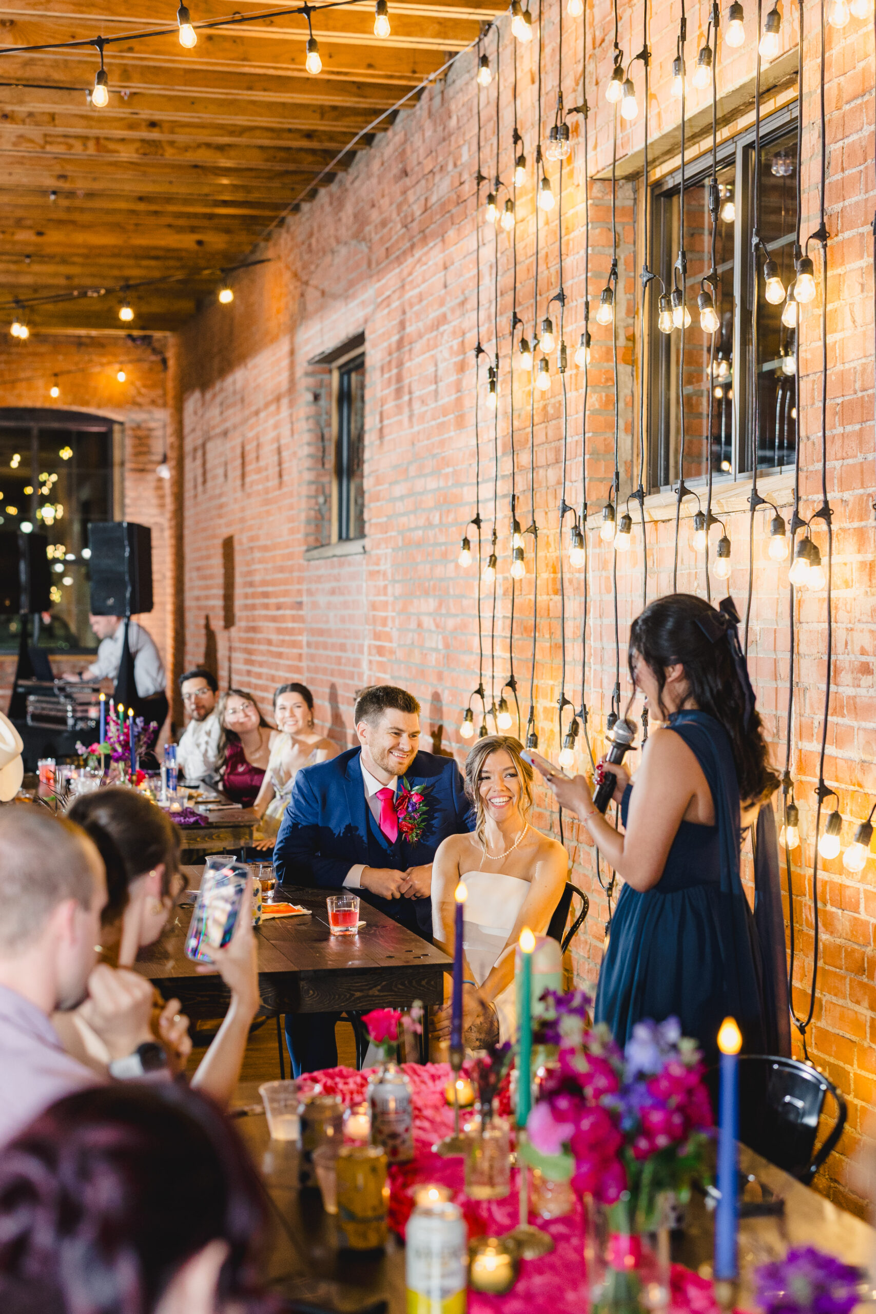 a couple sitting at a table in union in kansas city at their wedding reception next to a brick wall with string lights while one of their wedding guests gives a speech 