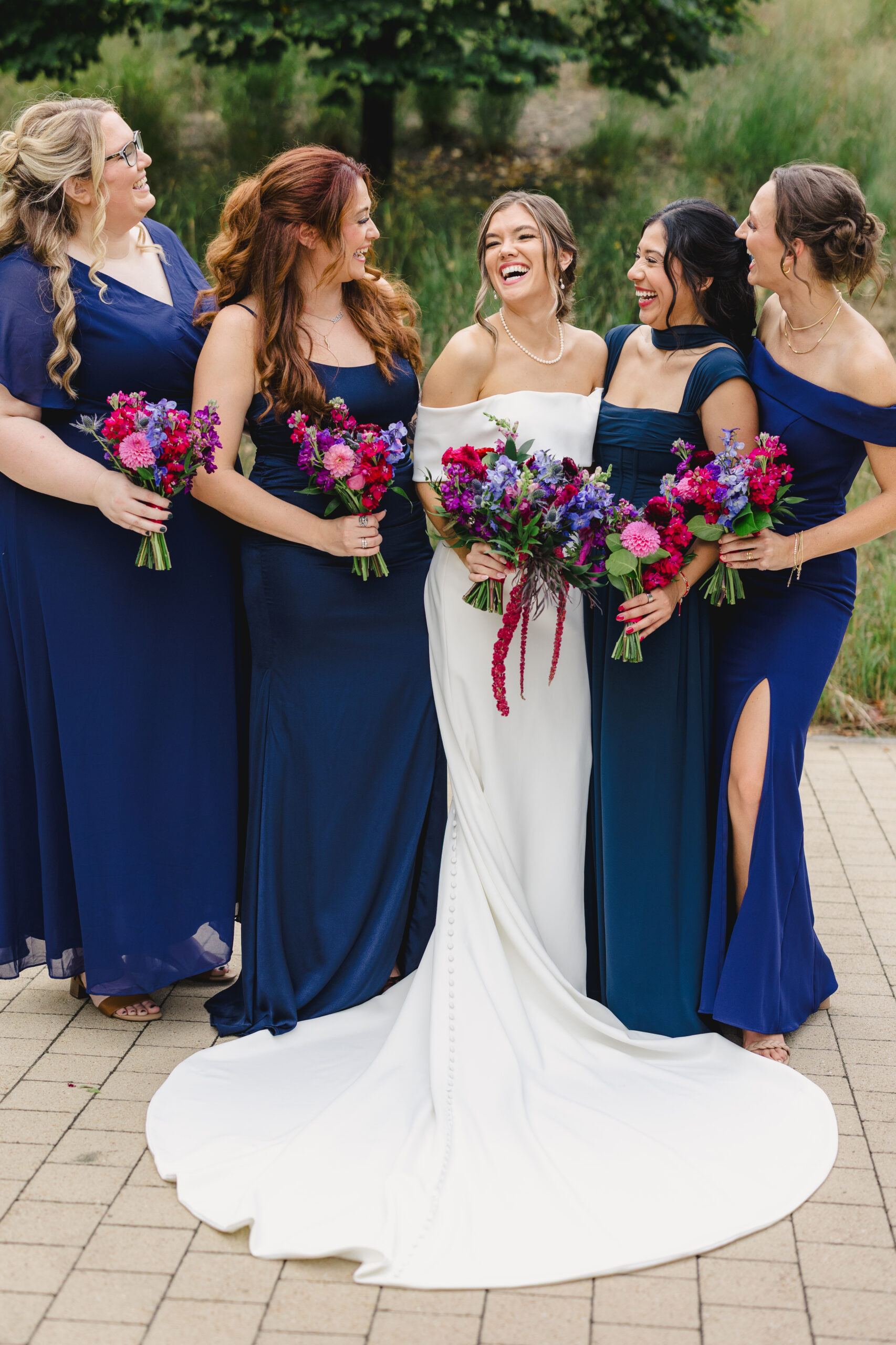a woman in a white wedding dress laughing with her bridal party who are all wearing blue dresses they are all holding matching bouquets in their hands consisting of blue, red and pink flowers 