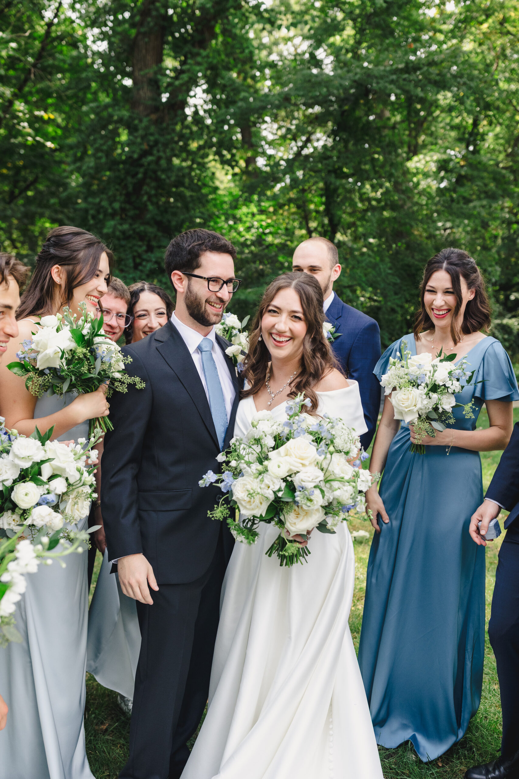 a couple taking wedding portraits with their wedding party on a green lawn the women are all holding bouquets of flowers that consist of white and blue flowers 