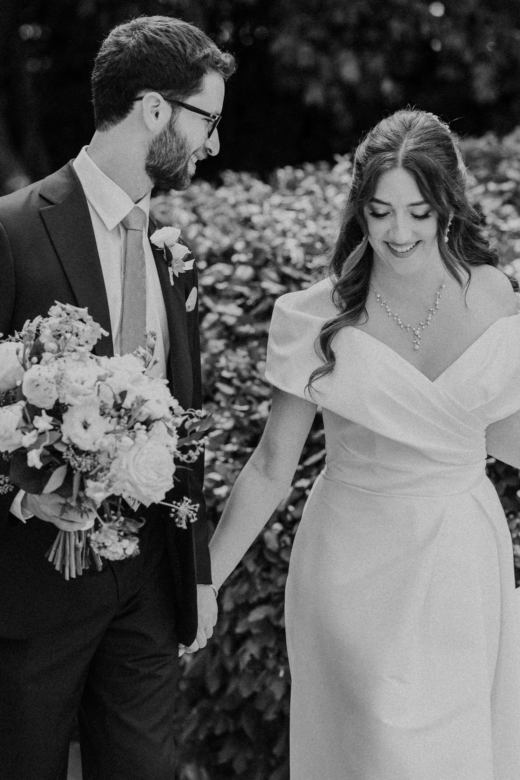 a couple holding hands and walking together the woman is wearing a white wedding dress and the man is wearing a black suit and holding her bouquet of flowers