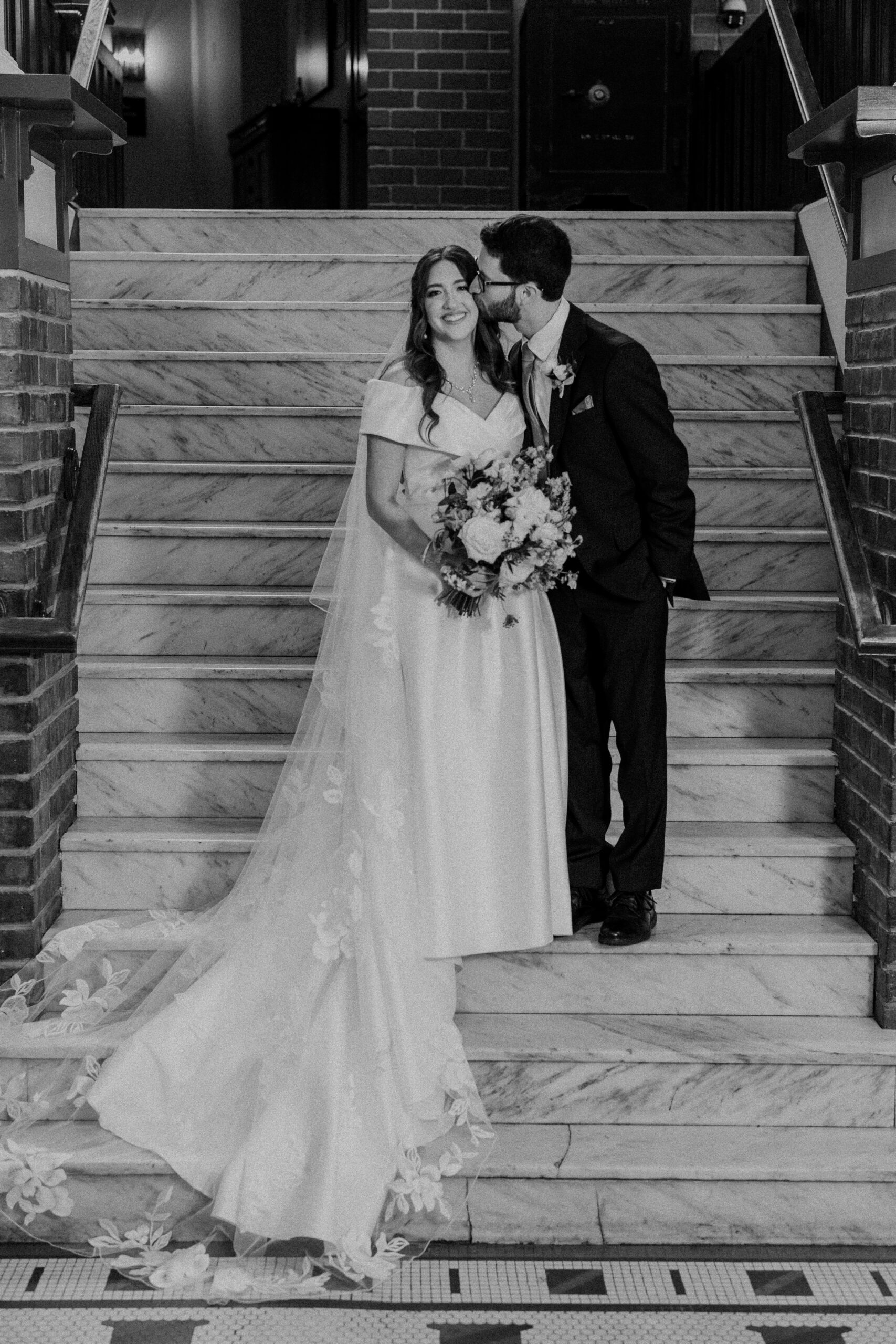 a couple taking wedding portraits on a staircase inside the elms hotel in kansas city the woman is holding a bouquet of flowers and the man is kissing her on her cheek