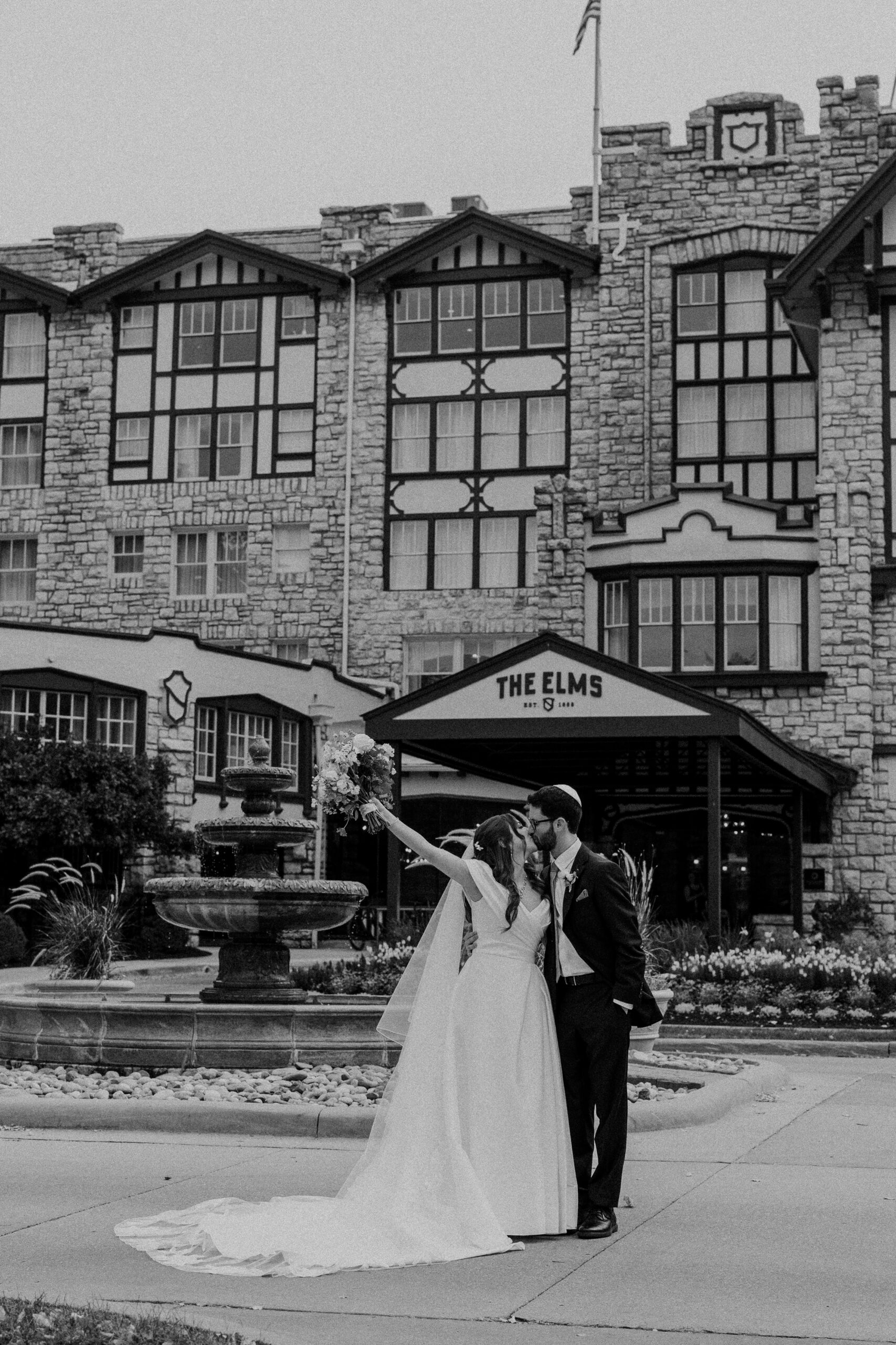 a couple taking wedding portraits in front of the elms hotel in kansas city they are kissing one another and the woman has one hand up in the air behind her and is holding a bouquet of flowers the woman is wearing a white wedding dress and veil the man is waering a black suit and is wearing a kippah