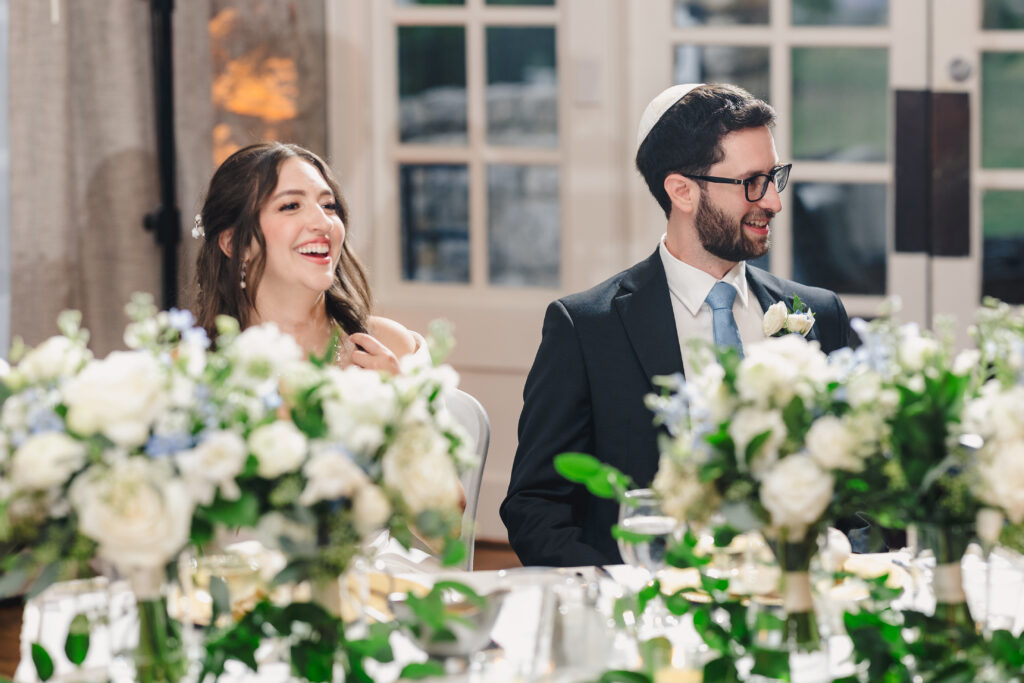 a couple sitting together at their wedding table at their wedding reception laughing in front of them are white and blue floral arrangements 
