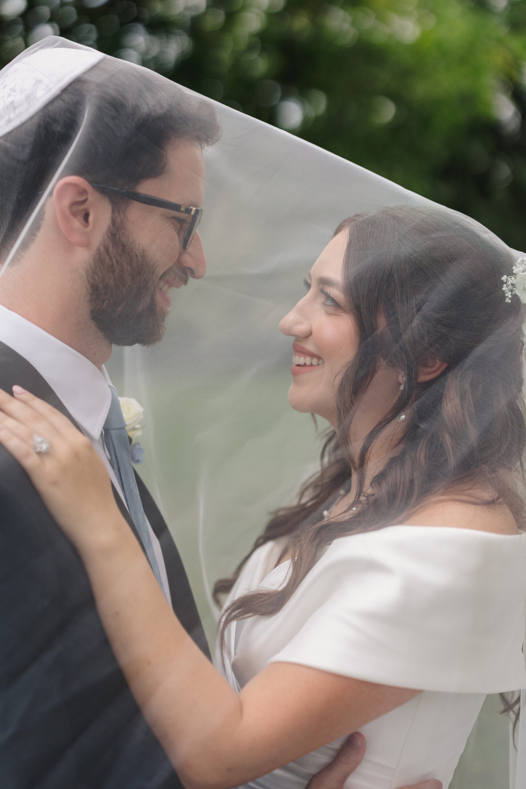 a couple taking couple wedding portraits they are both underneath of the bride's veil the bride is touching her partner's shoulder and they both smiling at each other the groom is wearing a kippah 