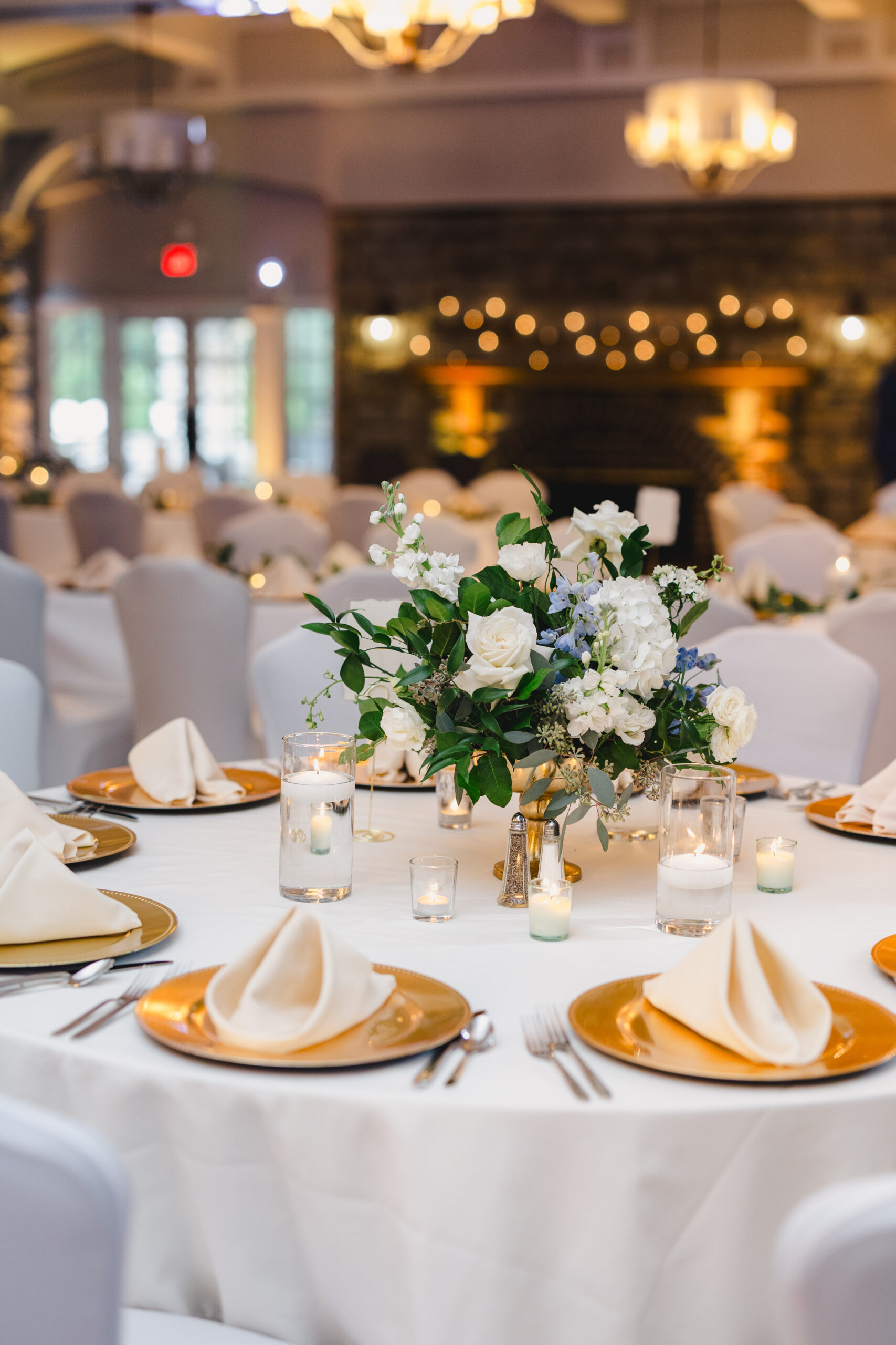 tables decorated with white table cloths, blue and white floral arrangements, gold plates, and candles inside of the reception room at the elms hotel