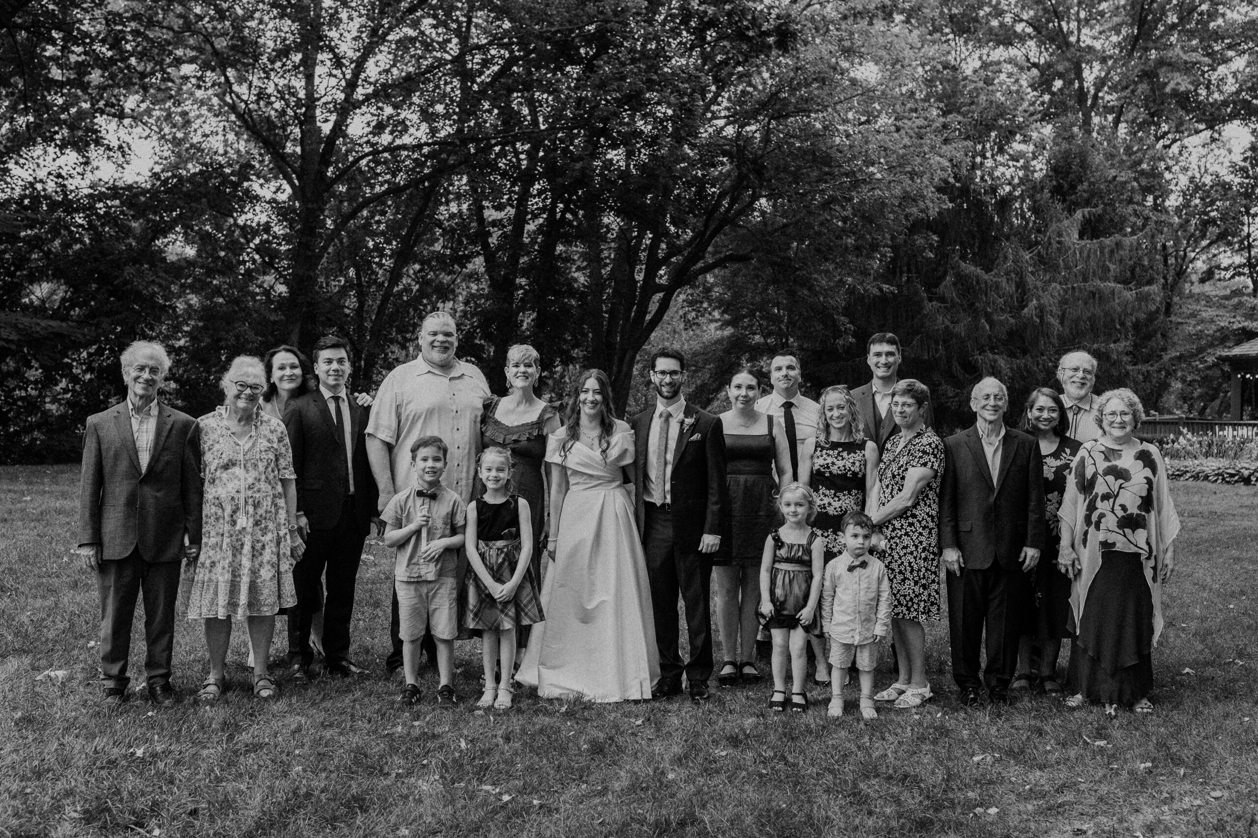 a bride and groom taking family wedding portraits on a green lawn in front of a cluster of trees 