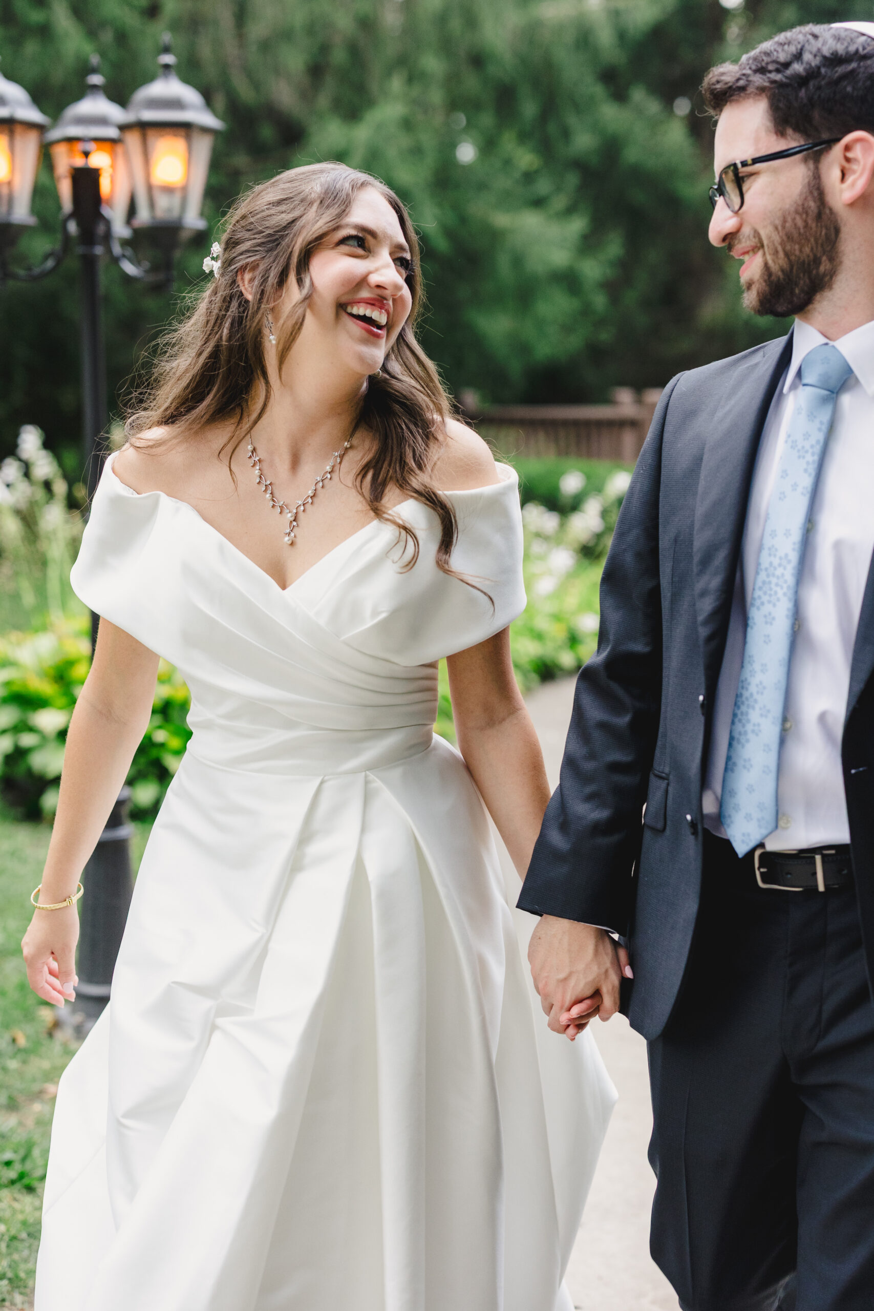 a couple walking hand in hand taking couple wedding portraits the woman is wearing a white wedding dress and laughing the groom is wearing a black suit they are both holding hands and smiling at each other 
