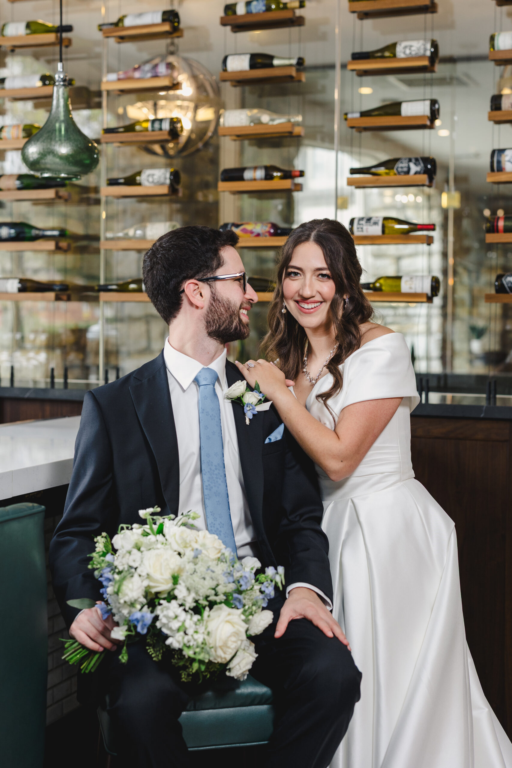 a couple taking couple wedding portraits inside of a bar room that is filled with wine bottles the man is sitting on a stool and holding the woman's bouquet and he is looking at her and smiling the woman is touching his shoulder and smiling 