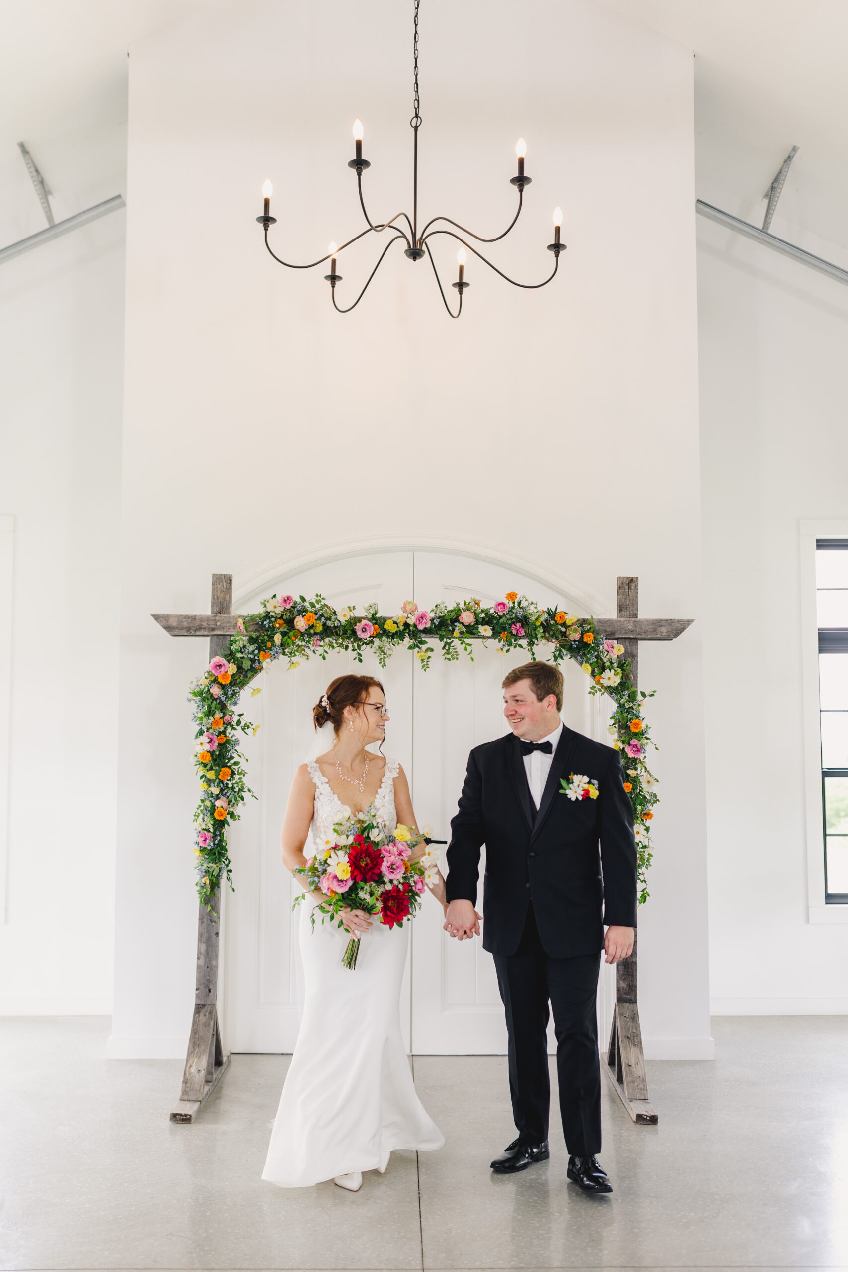 a couple holding hands in front of an altar decorated with flowers and looking at each other and smiling while a chandelier hangs above them 