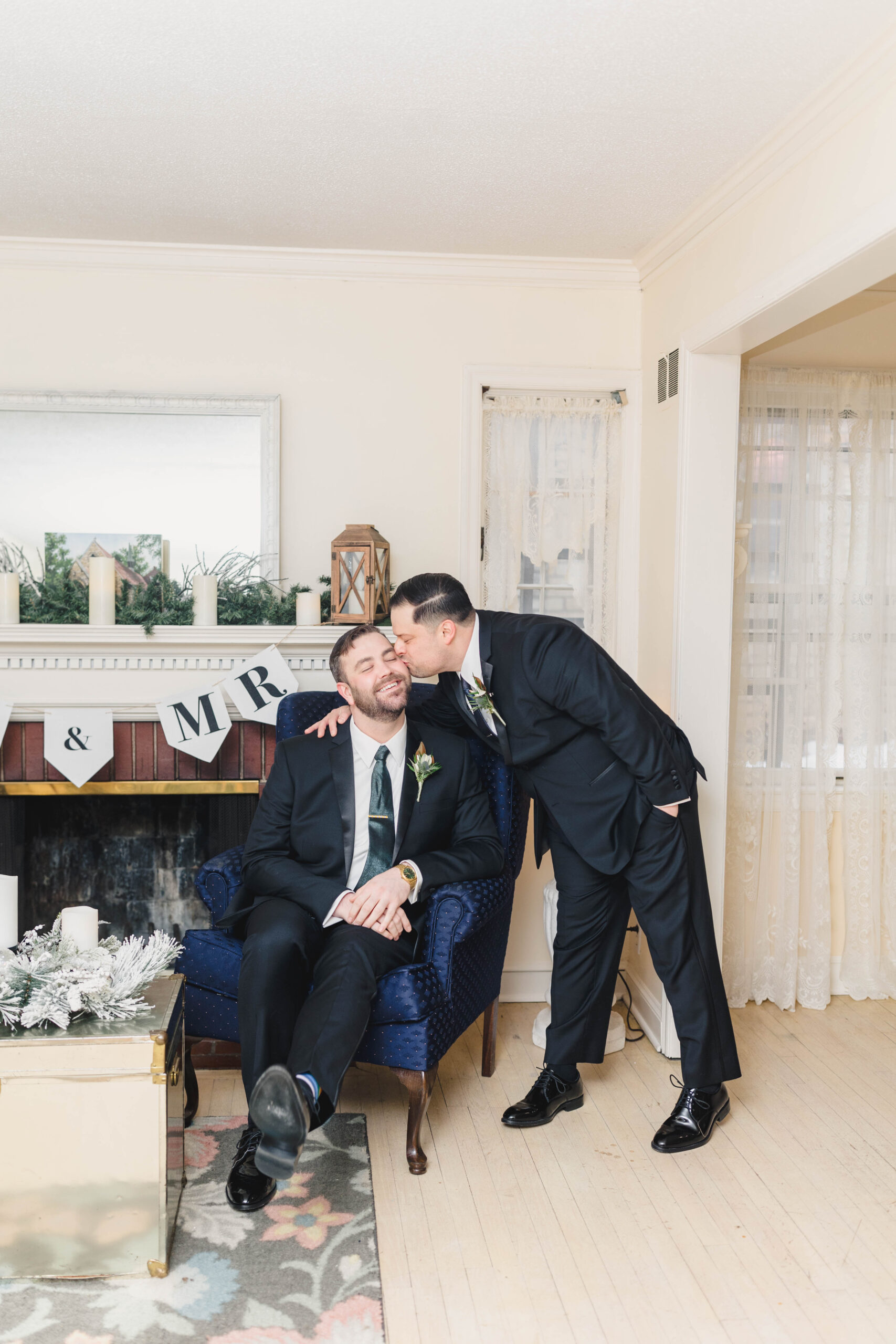 two men on their wedding day in front of a fireplace one groom is kissing the other groom on the cheek 