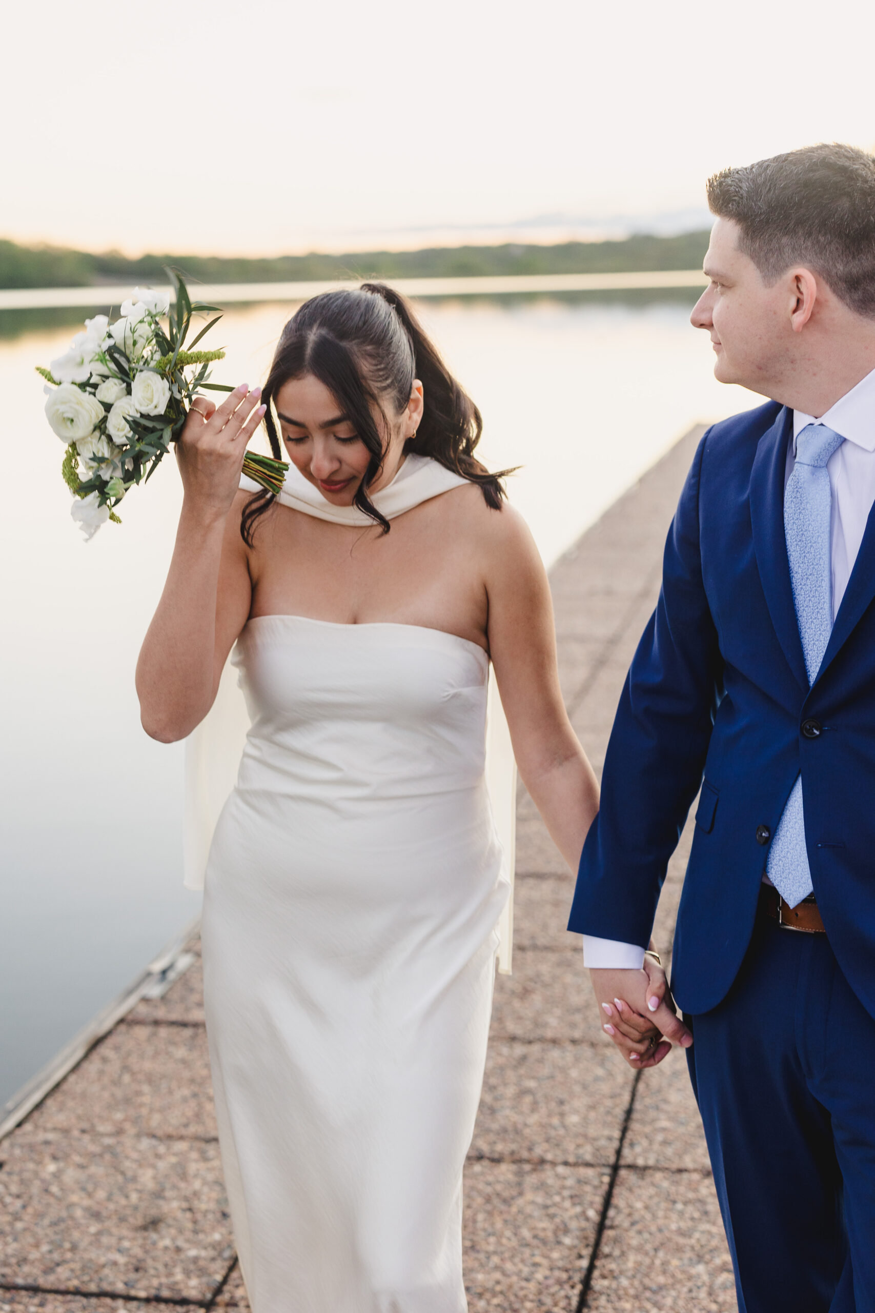 a bride and groom walking hand in hand down a sidewalk next to the lake the bride is fixing her hair 