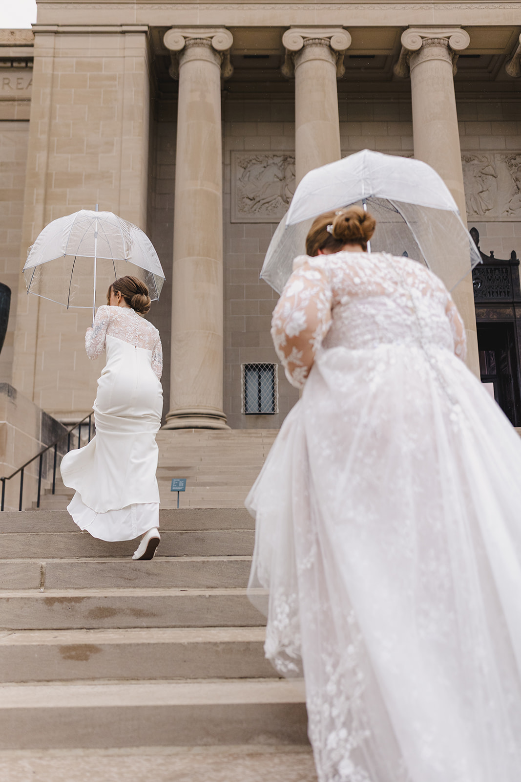 two brides walking up the staircase to the nelson atkins museum holding clear plastic umbrellas 
