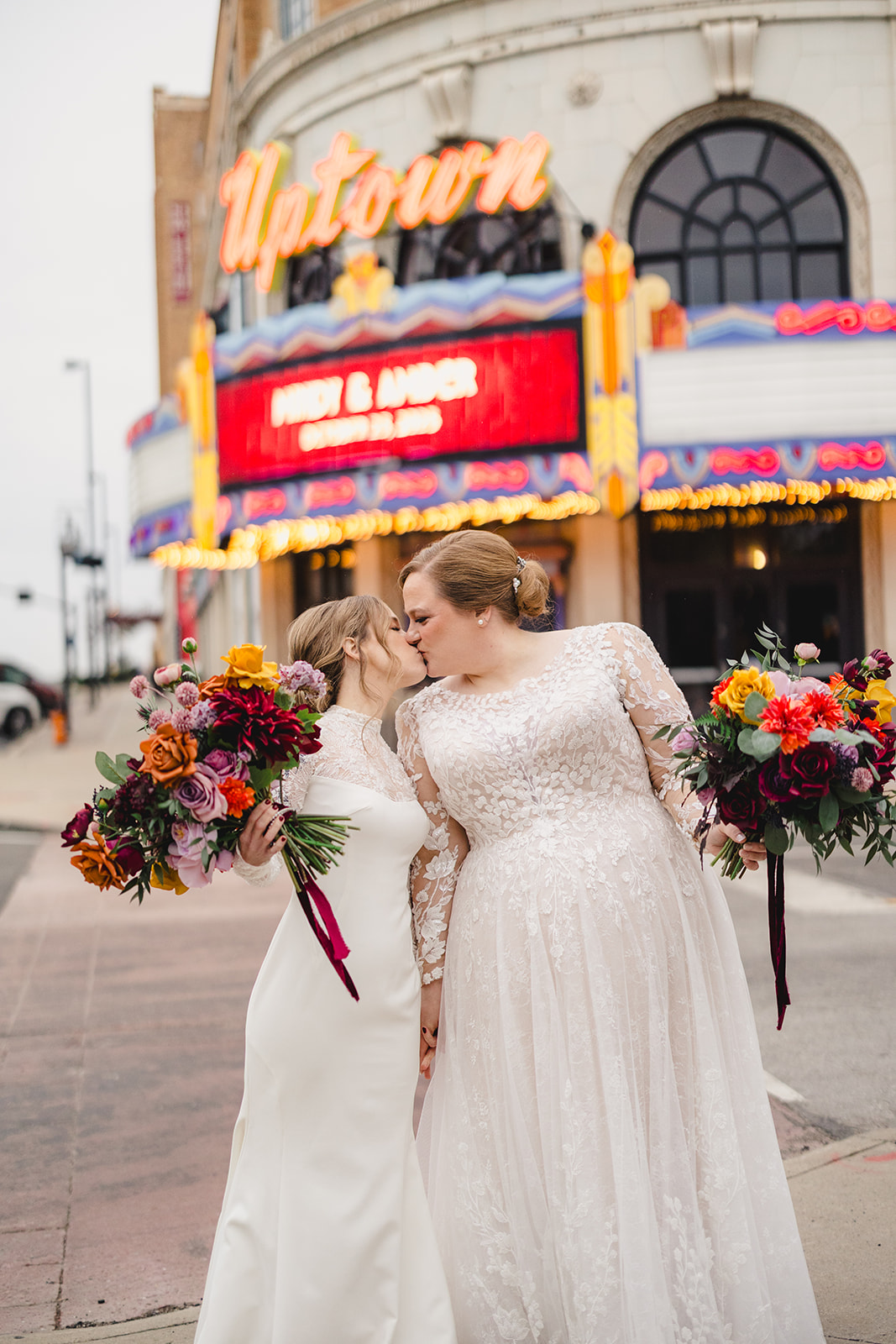 two brides holding floral bouquets and kissing in front of uptown theater in kansas city