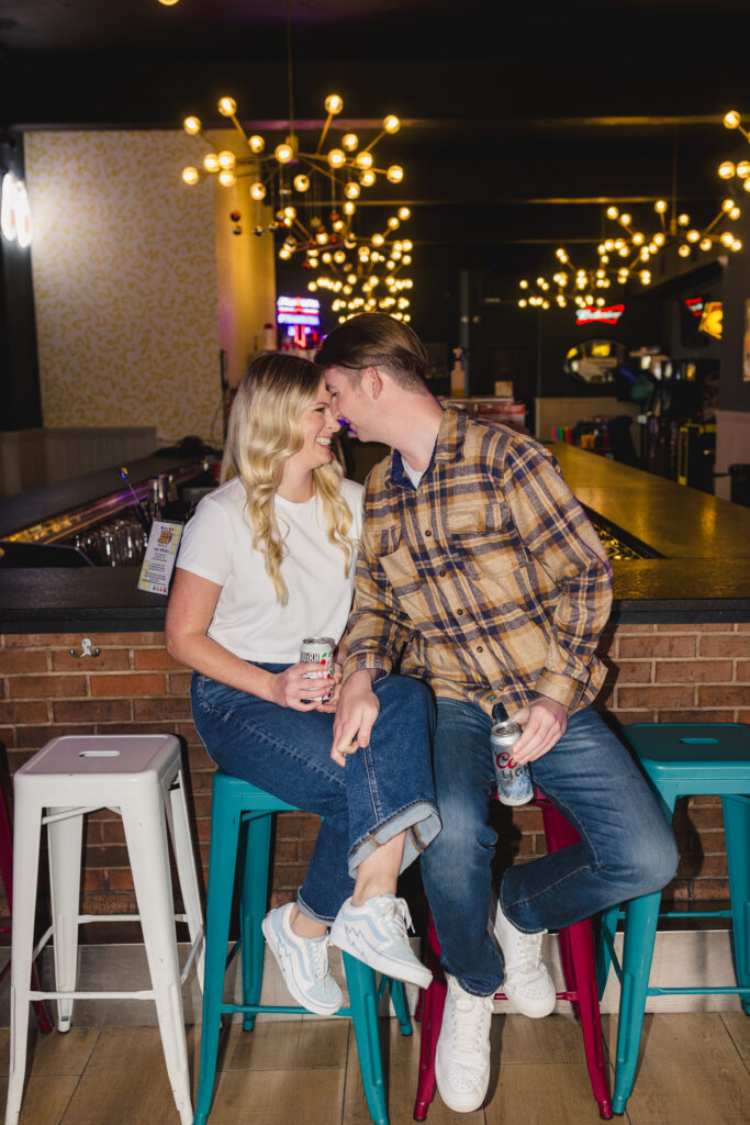 a man leaning towards his partner who is sitting on a barstool they are both smiling at each other and holding beers 