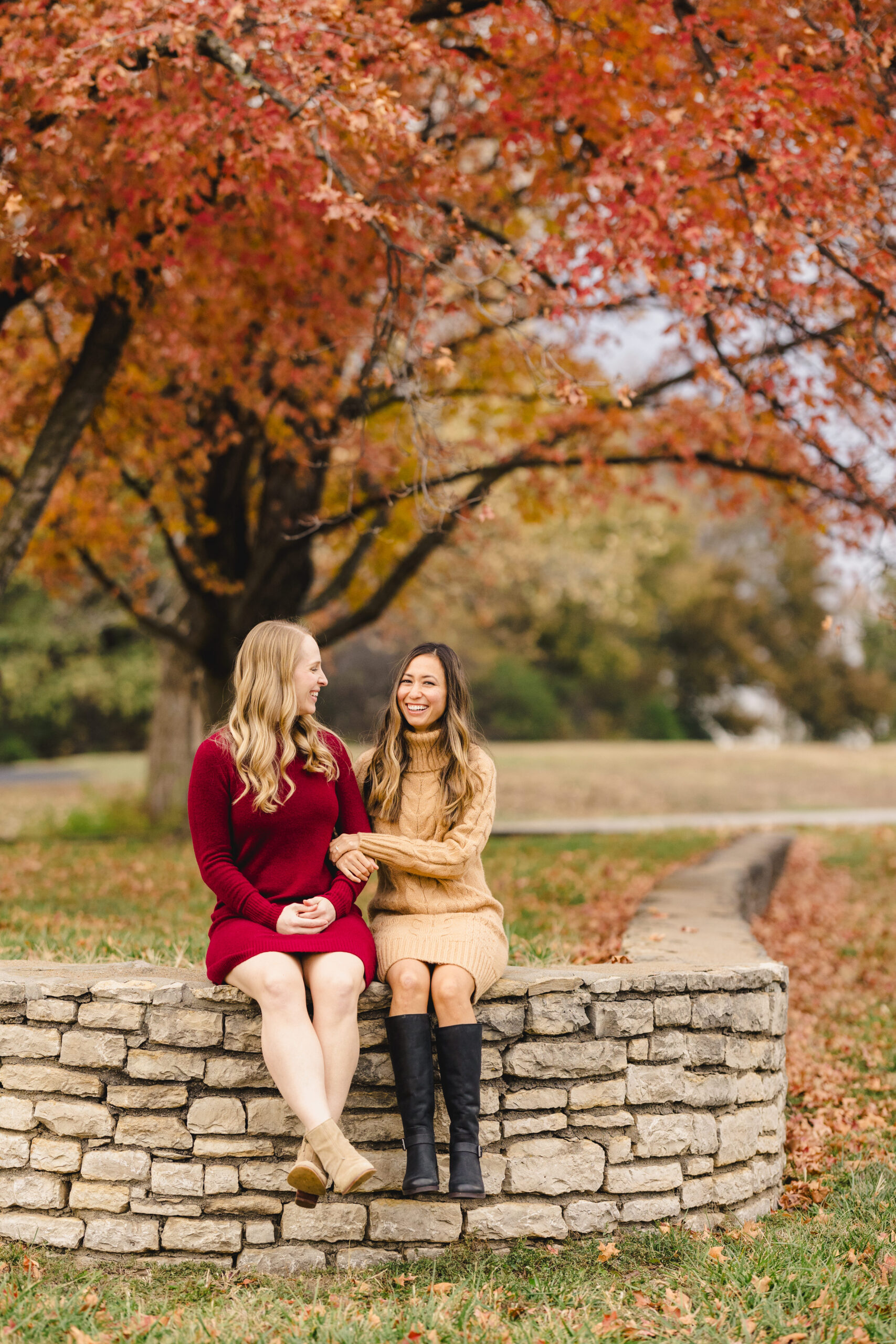two women sitting on a stone wall in a park arm in arm 